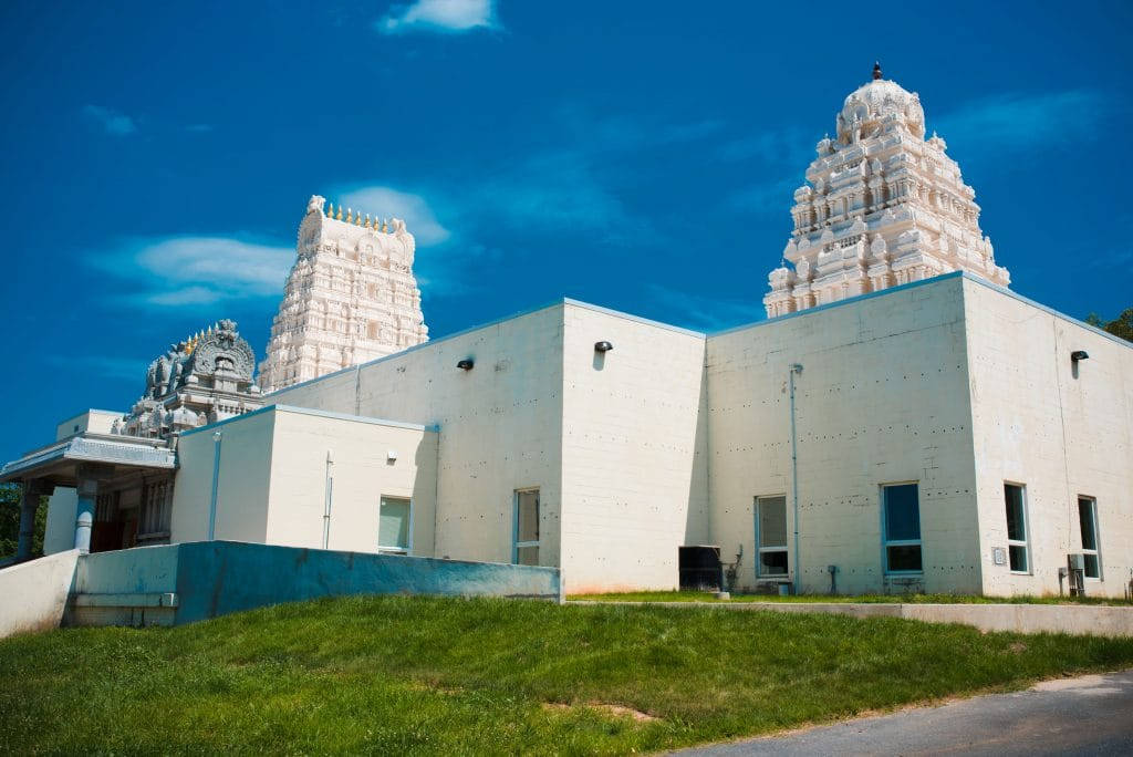 Hindu Temple 桌布 壁紙 — 一座白色寺廟擁有華麗的塔樓，矗立在現代混凝土建築旁，天空湛藍，雲朵稀疏。