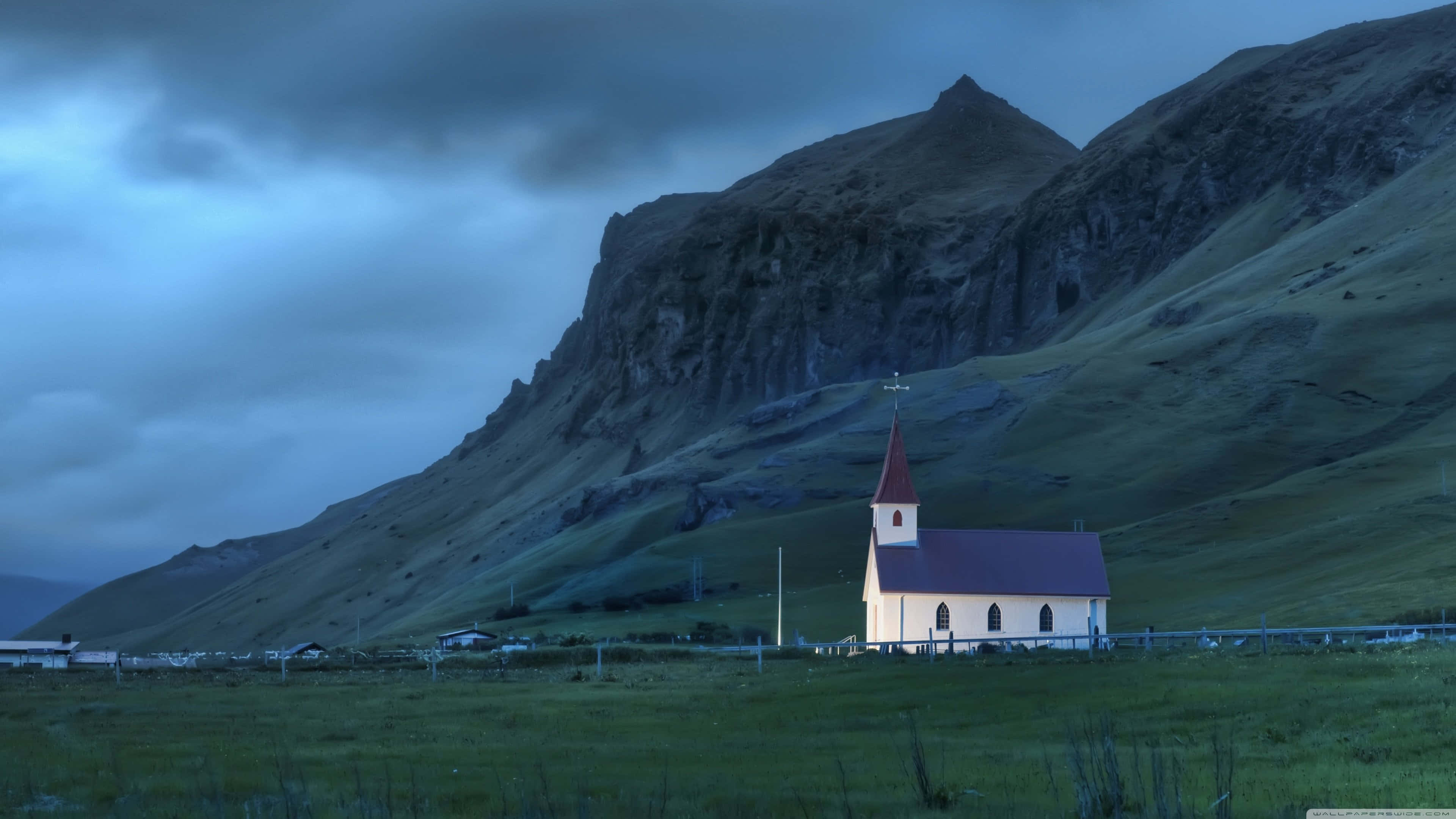 Iceland Desktop 桌布 壁紙 — 一座白色小教堂，紅色屋頂，孤獨地矗立在廣闊的綠色田野中，陰沉的暮色天空下，群山巍峨。