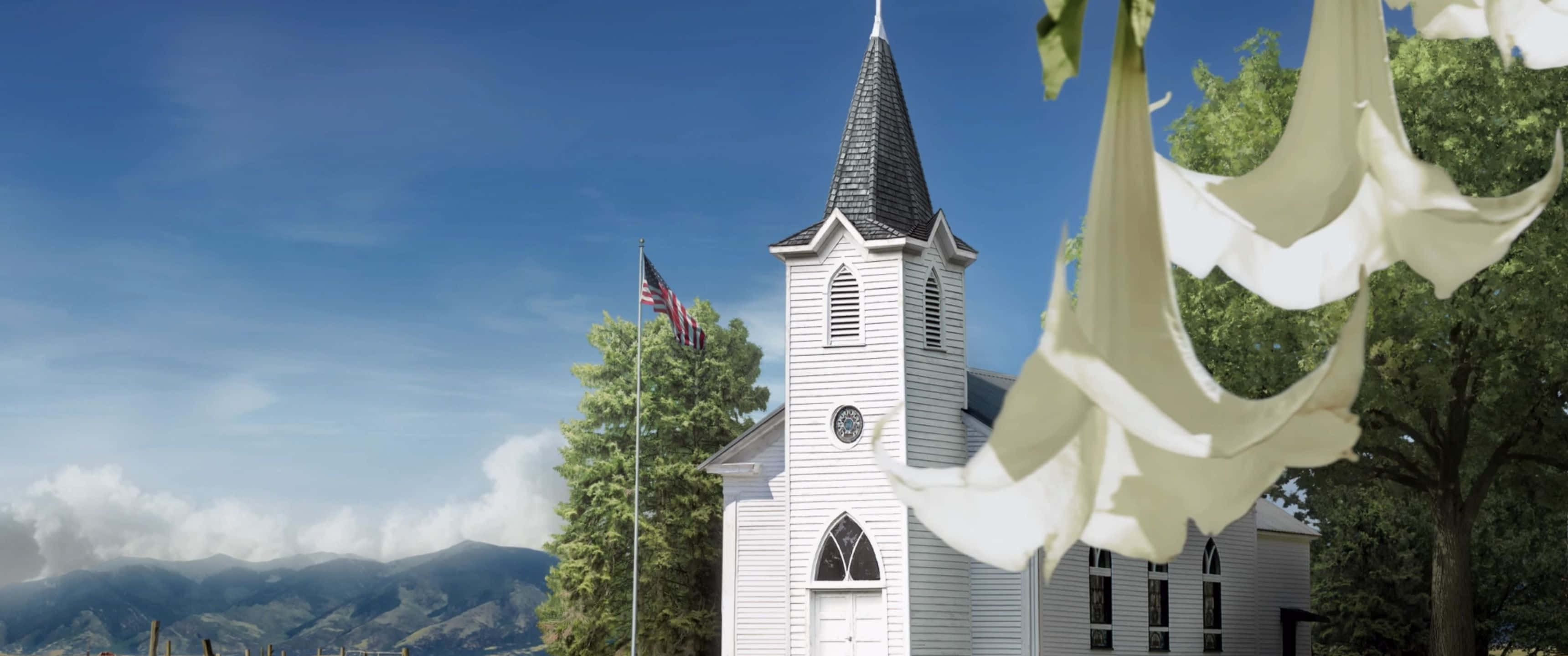 3440x1440p Far Cry 5 桌布 壁紙 — 一座白色教堂帶有尖頂，矗立在藍天下，遠方有山巒，前景有白色花朵。
