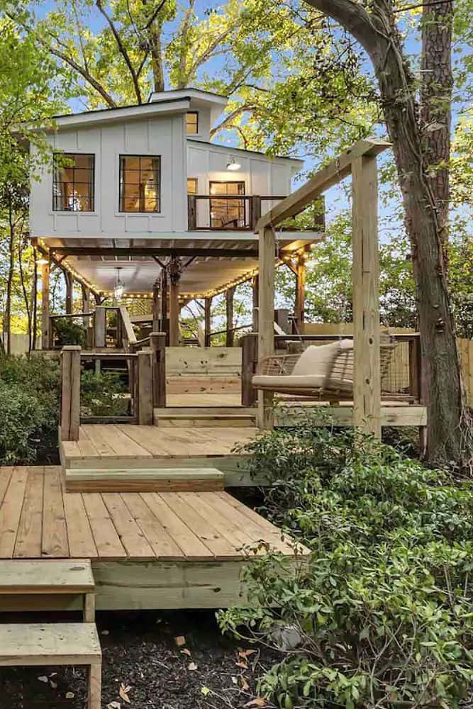 Cottage 桌布 壁紙 — 一座白色樹屋，設有木製平台和串燈，坐落在高大的綠樹之間，在清澈的藍天下。