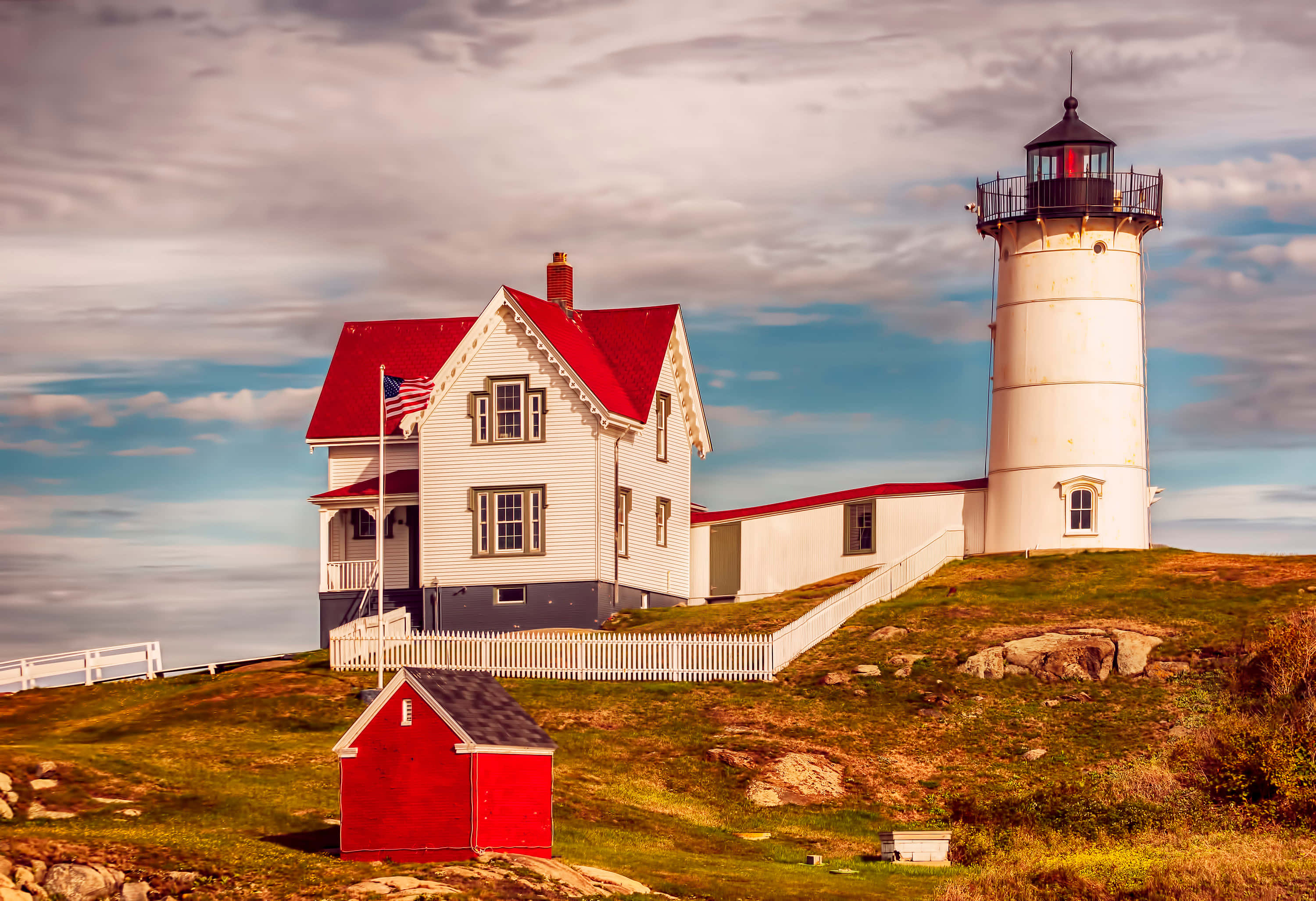 Lighthouse 桌布 壁紙 — 一座白色燈塔和紅色屋頂的管理員小屋矗立在草叢生的懸崖上，俯瞰著多雲天空下的大海。