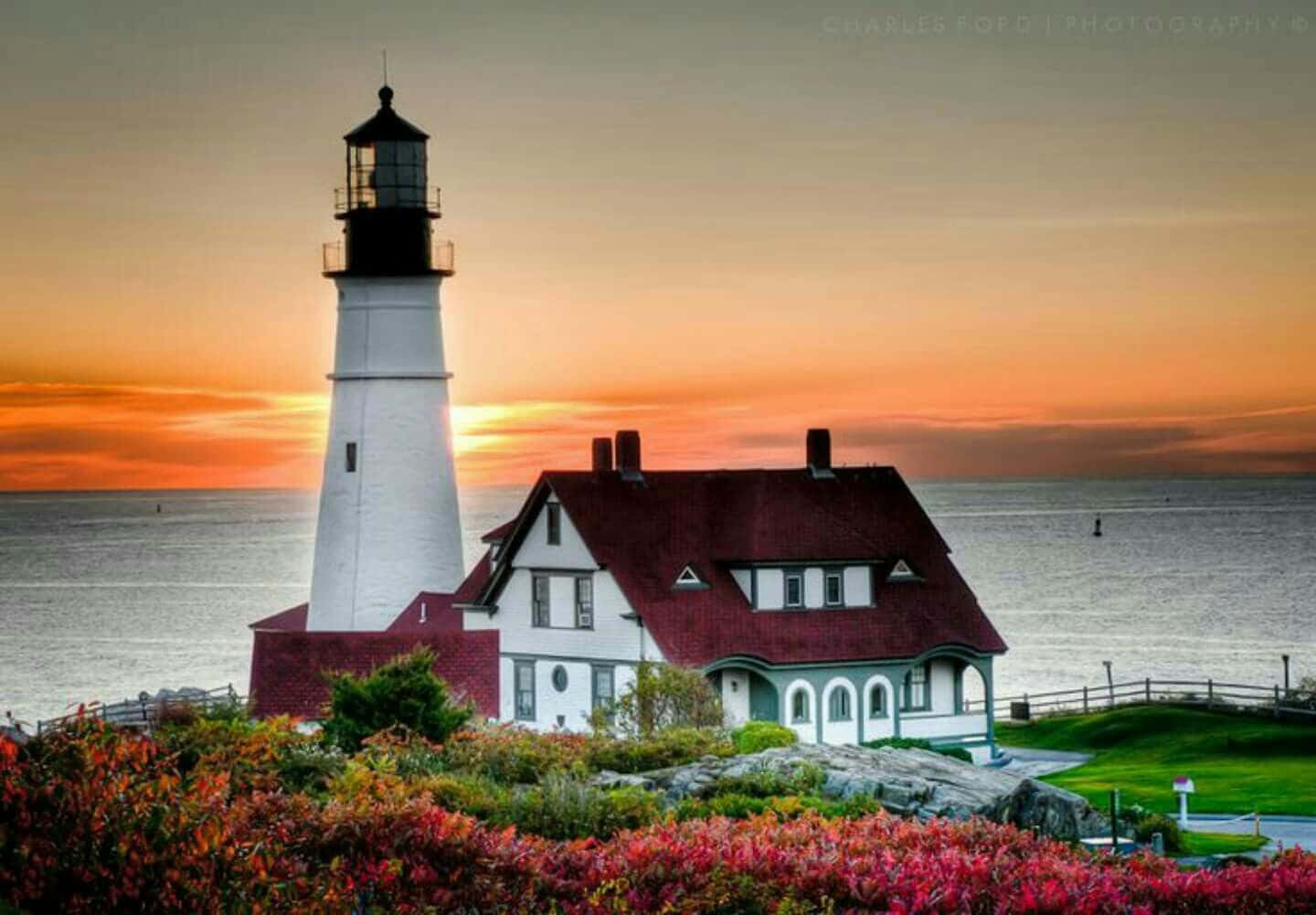 Lighthouse 桌布 壁紙 — 一座白色燈塔和紅頂看守員小屋矗立在懸崖上，夕陽西下，天空火紅橙黃，下方海洋平靜。