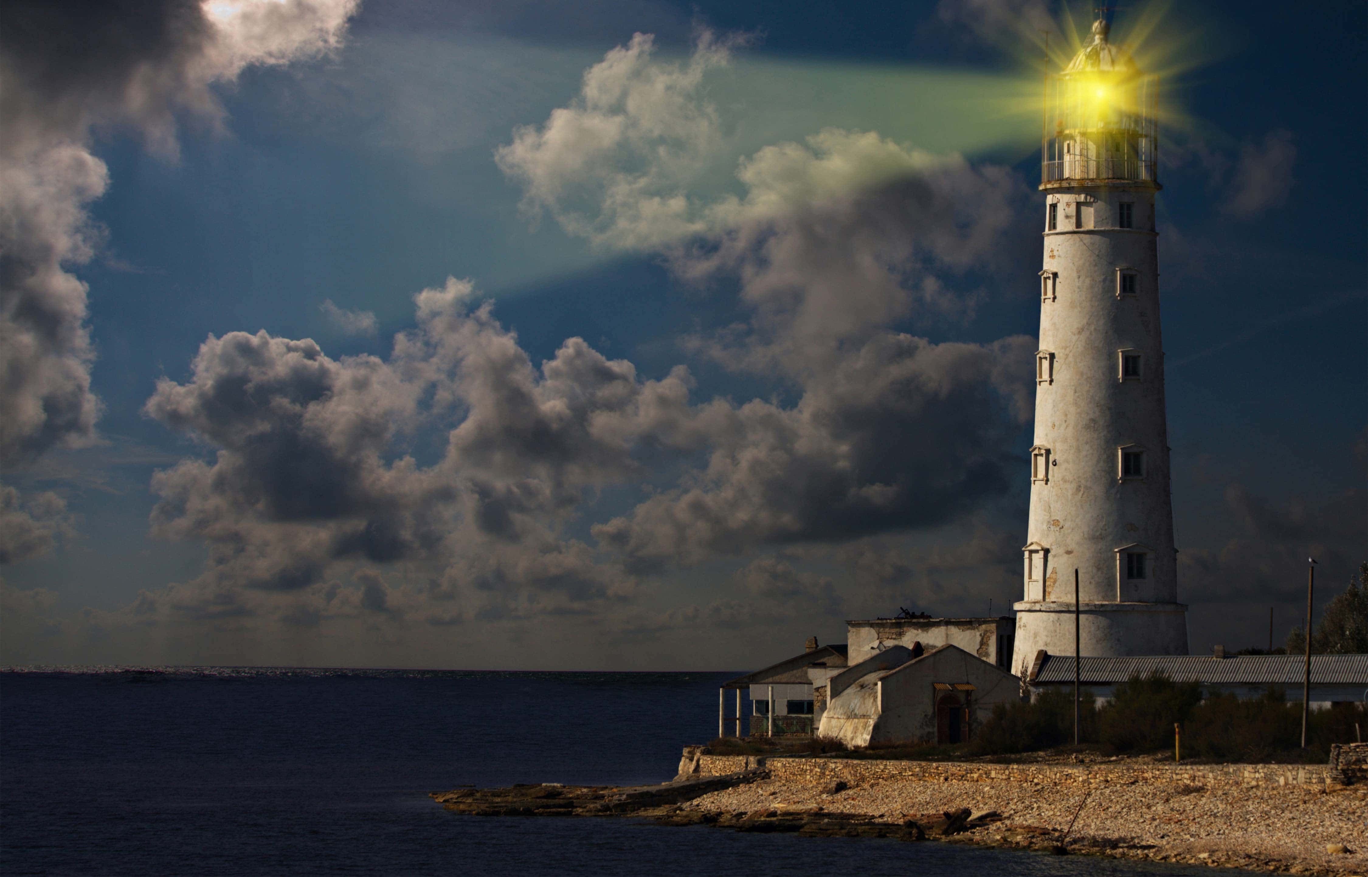 Lighthouse 桌布 壁紙 — 一座白色燈塔在黃昏時分，穿過戲劇性的雲層，向平靜的海面發出明亮的黃色光芒。