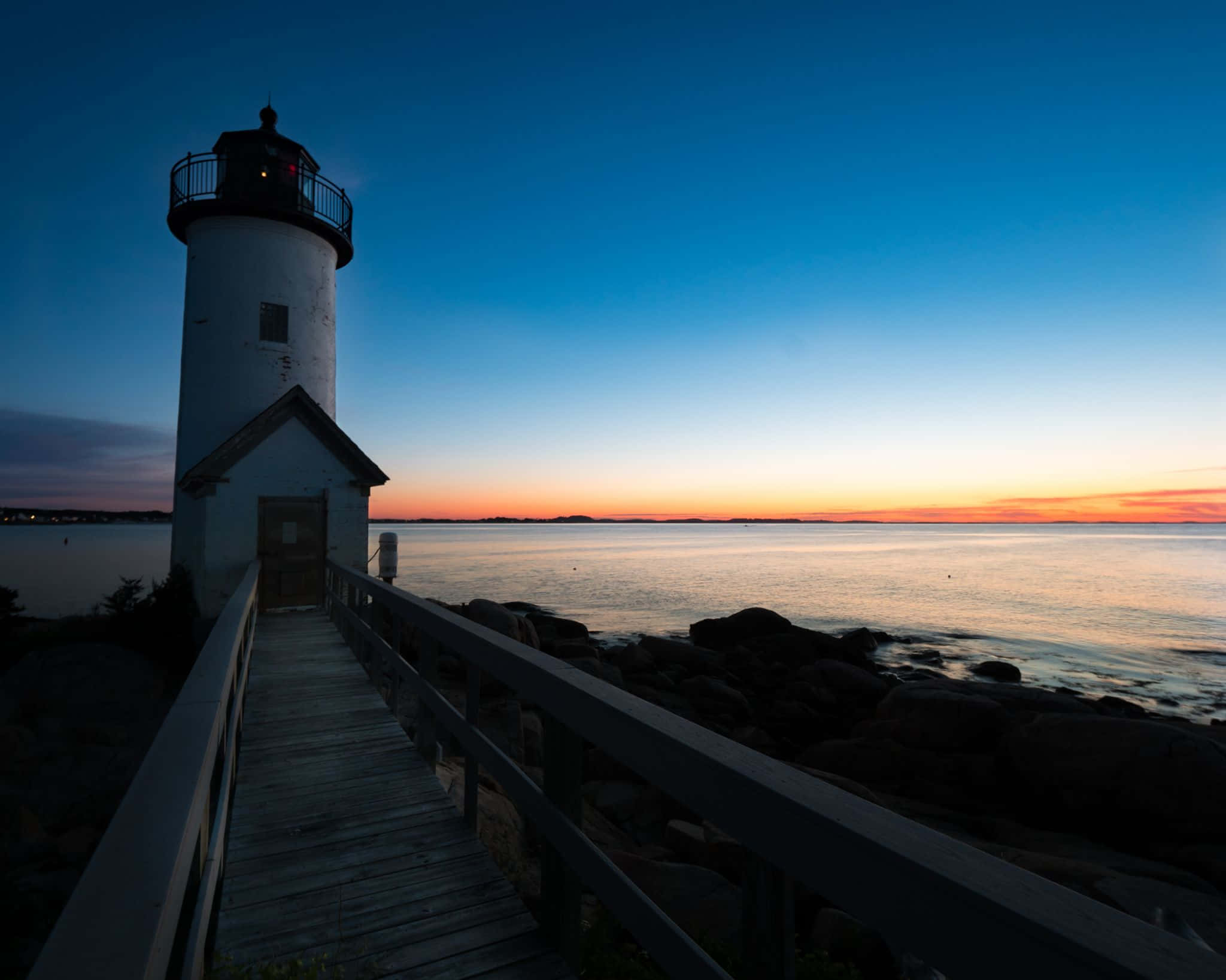 Lighthouse 桌布 壁紙 — 一座白色燈塔在黃昏時分矗立，一條木製步道通往燈塔，天空在平靜的水面上閃耀著橙色和藍色。