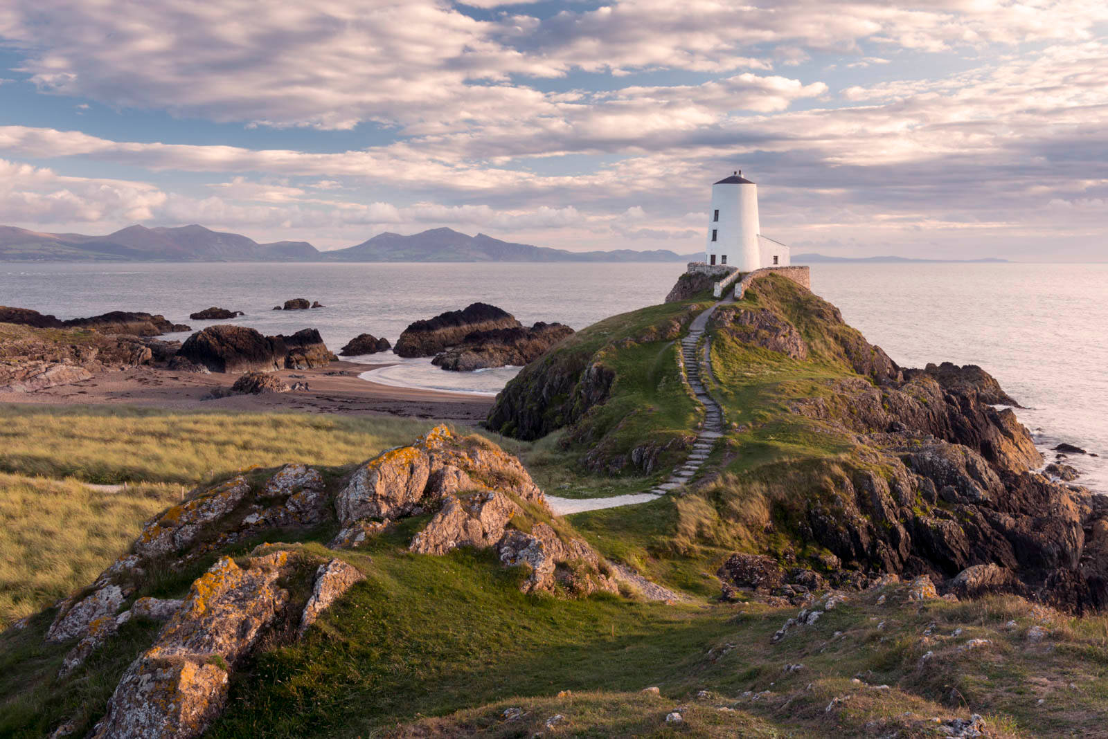 Lighthouse 桌布 壁紙 — 一座白色燈塔坐落在長滿草的懸崖上，俯瞰著平靜的大海和遠處的山脈，天空局部多雲。