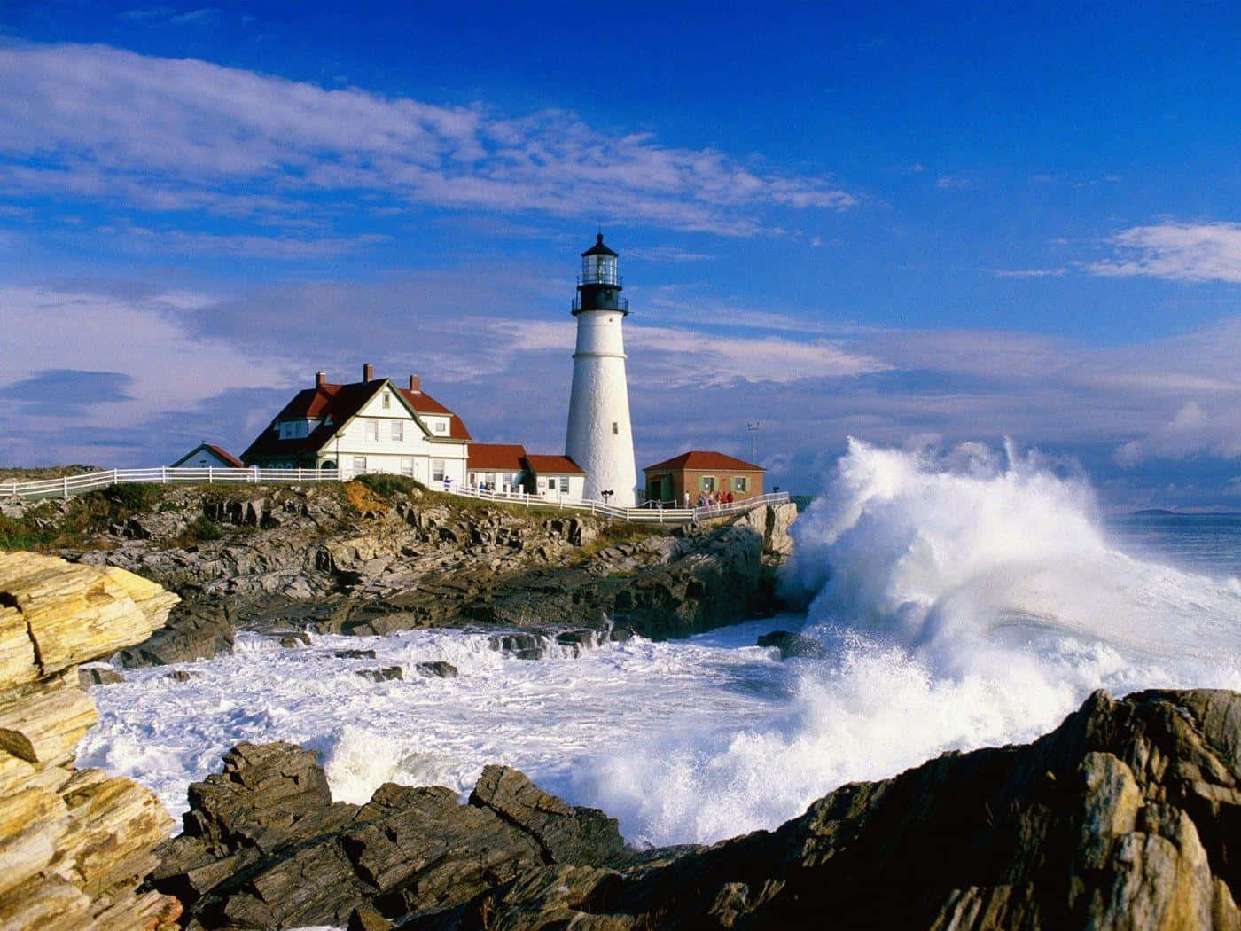 Lighthouse 桌布 壁紙 — 一座白色燈塔屹立在岩石峭壁上，海浪撞擊在蔚藍的天空下爆發出白色泡沫。