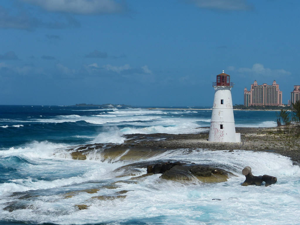 Nassau Bahamas 桌布 壁紙 — 一座白色燈塔屹立在岩石海岸上，強勁的海浪在明亮的藍天下拍打著它。