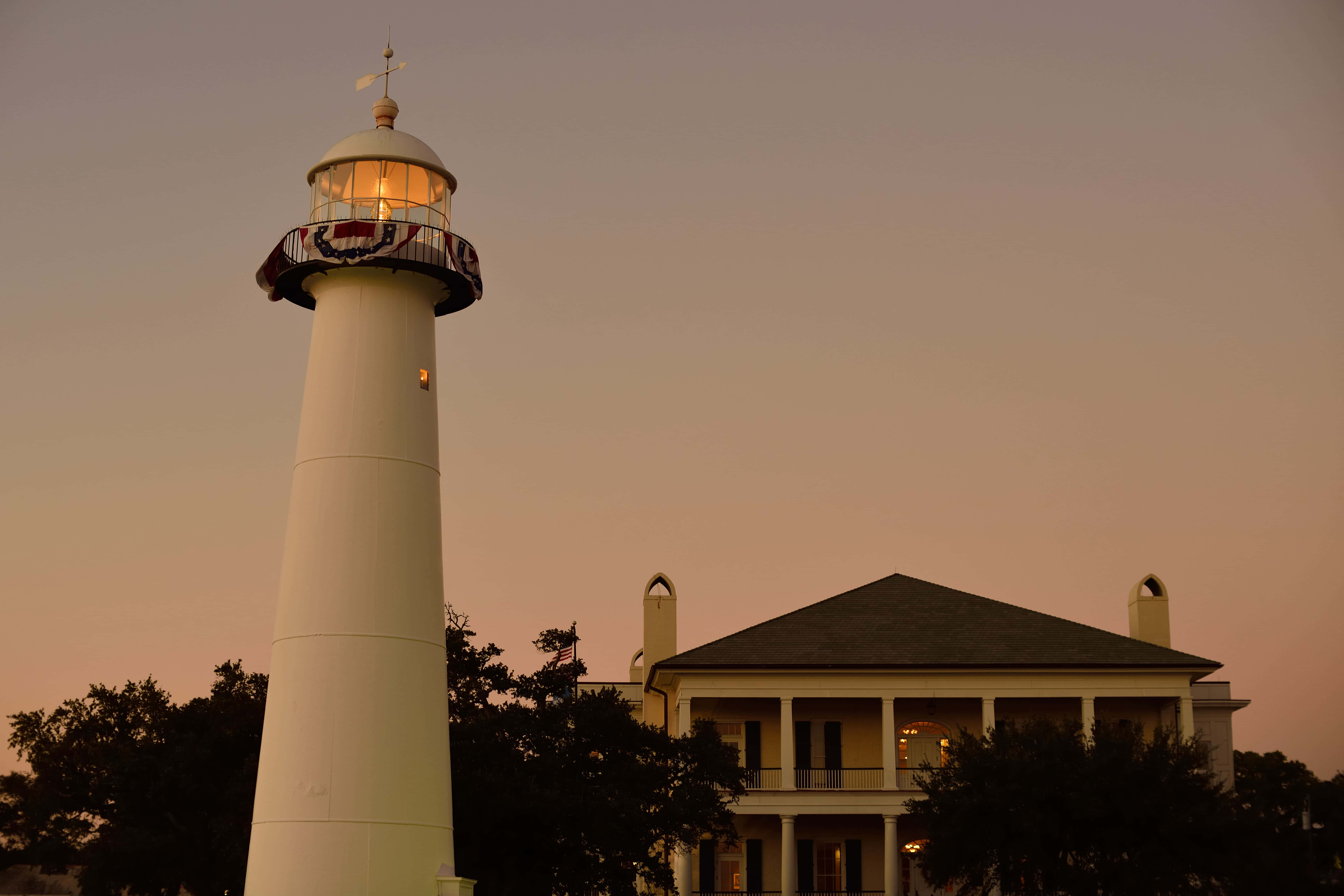 Lighthouse 桌布 壁紙 — 一座白色燈塔帶有發光的燈室，在黃昏時分站在一座殖民風格房屋旁邊，天空色調溫暖。