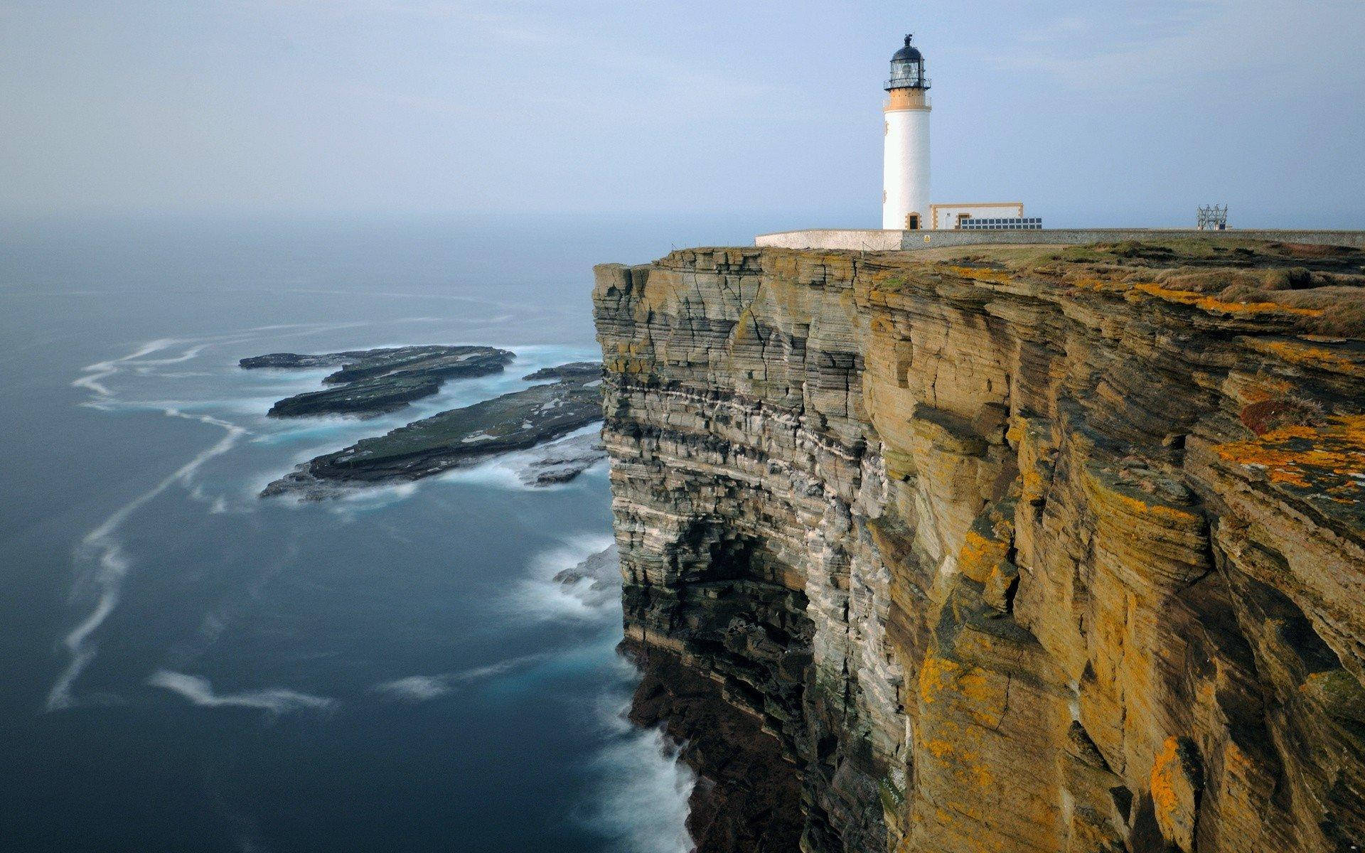 Lighthouse 桌布 壁紙 — 一座白色燈塔棲息在崎嶇的懸崖上，俯瞰著波濤洶湧的海浪拍打岩石海岸。