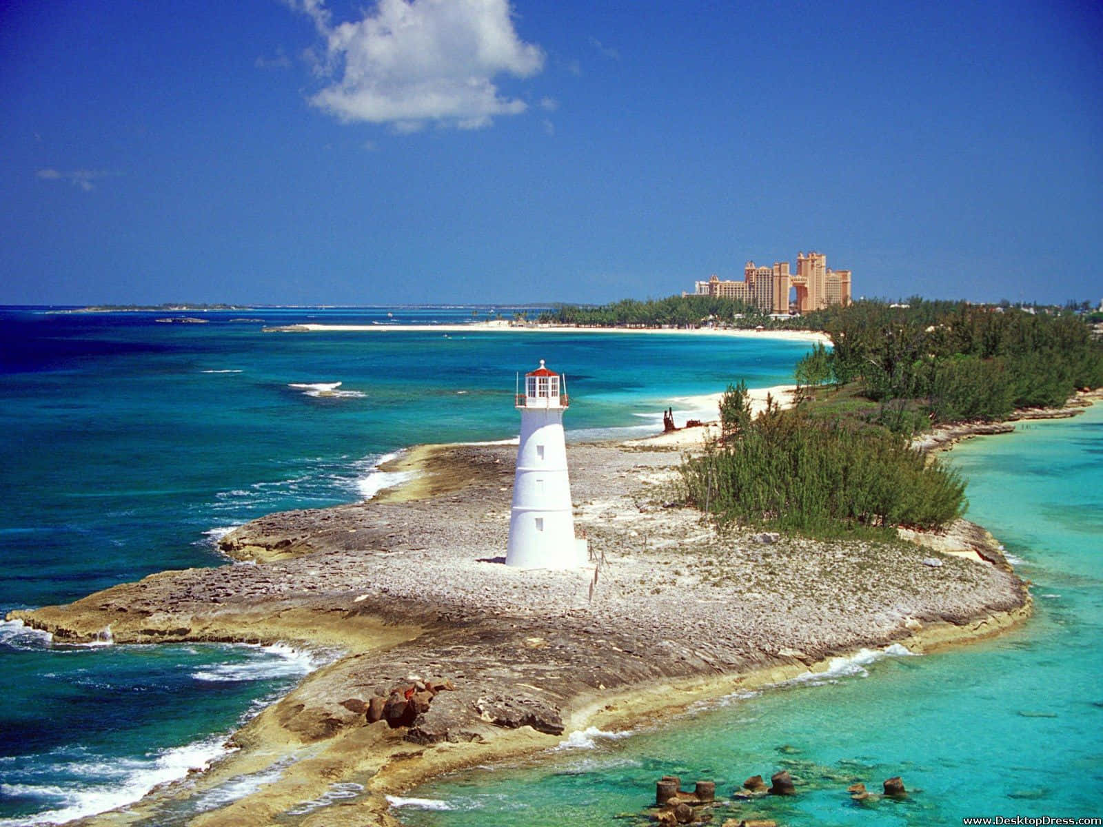 Bahamas Beach 桌布 壁紙 — 一座白色燈塔矗立在岩石島上，四周環繞著碧綠的海水，遠處的海岸上可以看到一個豪華度假村。