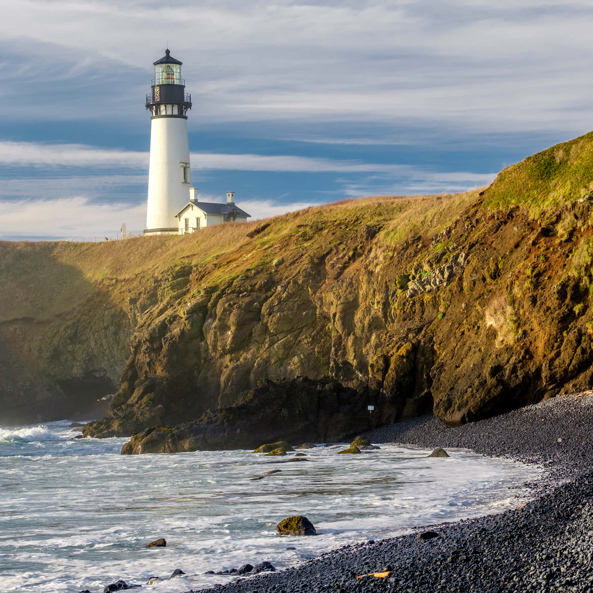 Oregon Coast 桌布 壁紙 — 一座白色燈塔矗立在崎嶇的懸崖頂上，俯瞰著在部分多雲的天空下拍打的海浪和卵石海灘。