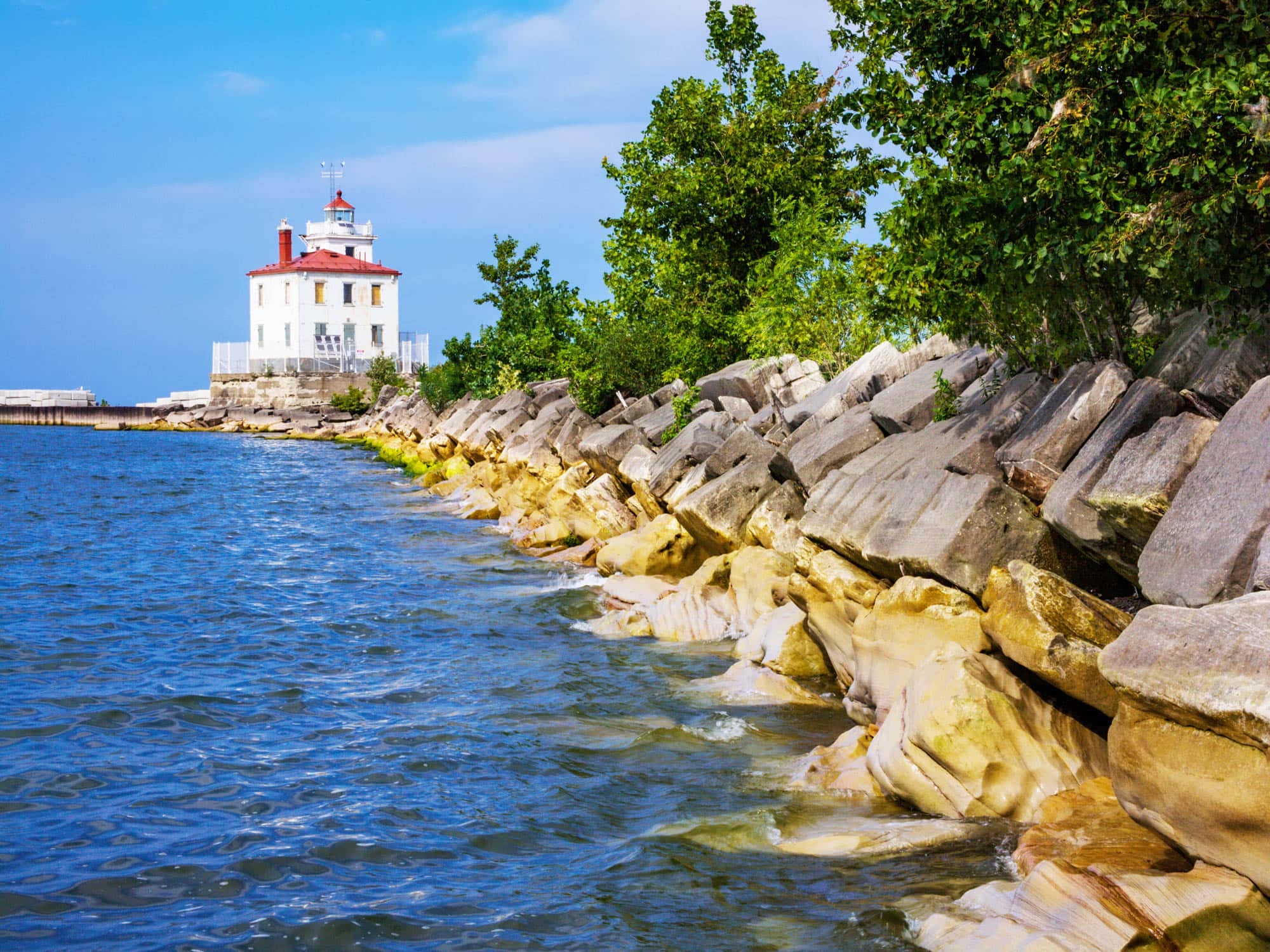 Lake Erie 桌布 壁紙 — 一座白色燈塔配紅色屋頂矗立在岩石海岸上，海浪拍打著風化的石頭，天空清澈湛藍。