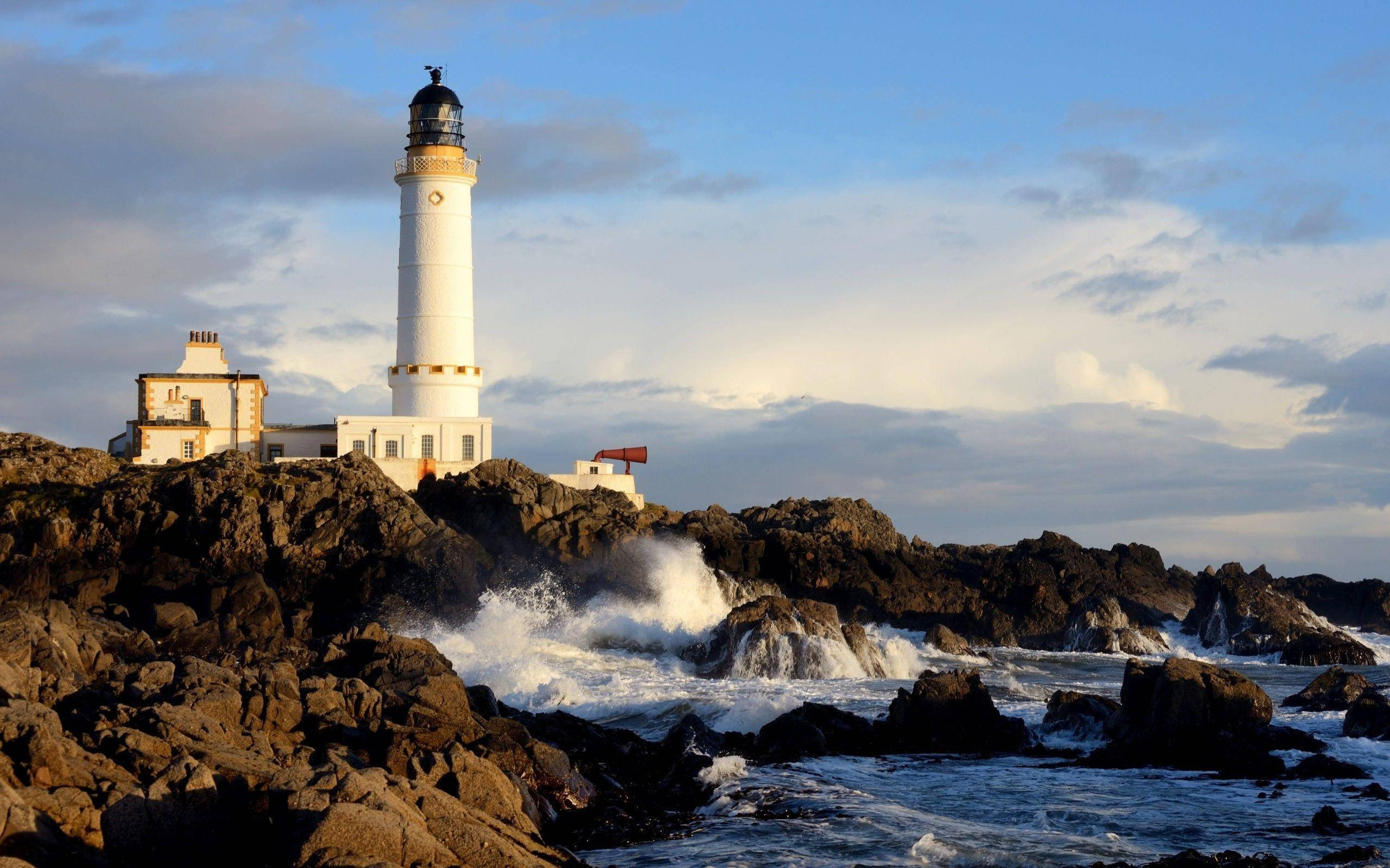 Lighthouse 桌布 壁紙 — 一座白色燈塔高聳在崎嶇的懸崖上，破碎的海浪在部分多雲的天空下拍打著黑色的岩石。