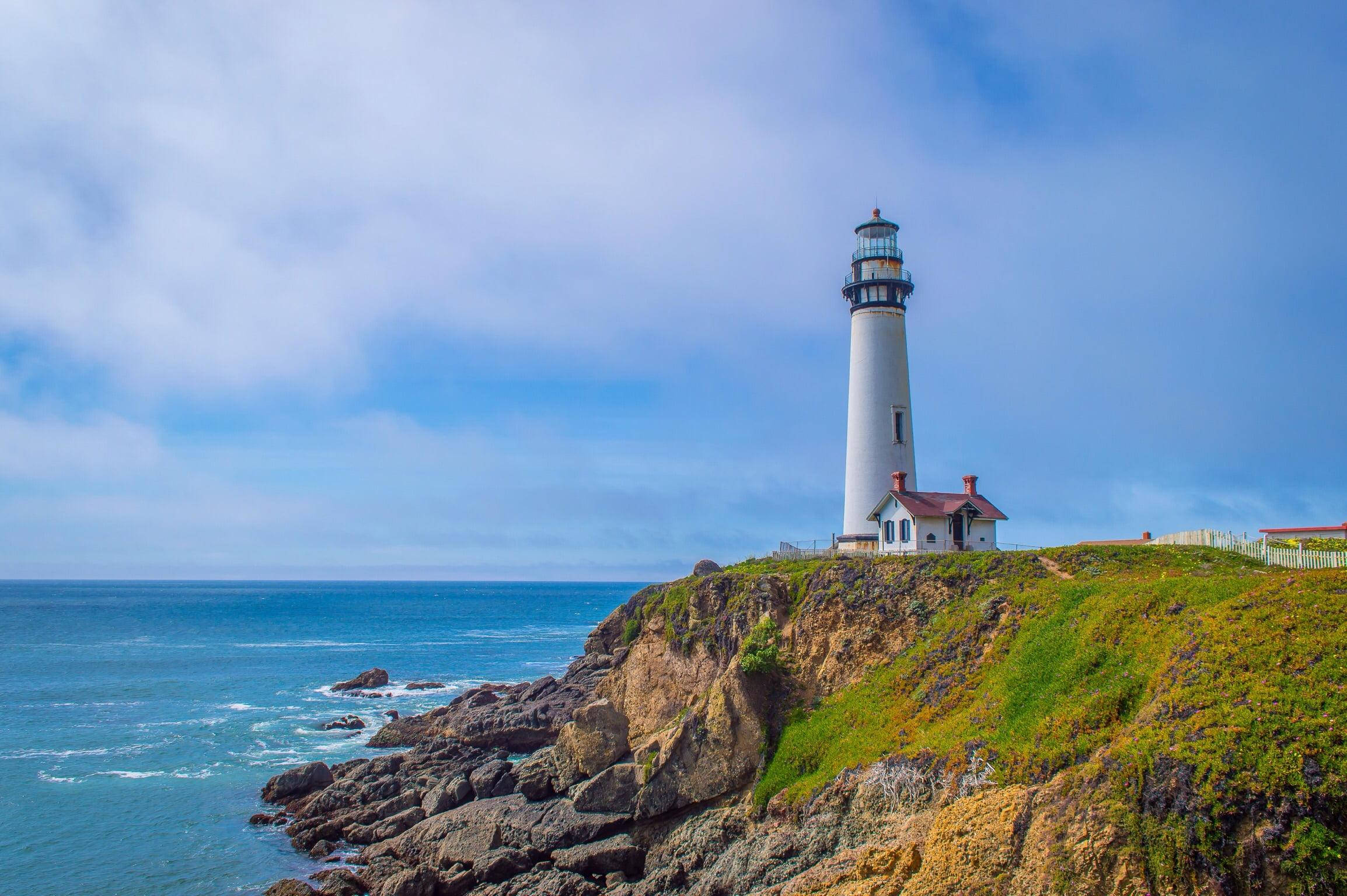 Lighthouse 桌布 壁紙 — 一座白色燈塔高高矗立在長滿青草的懸崖上，俯瞰著深藍色的海洋，天空多雲。
