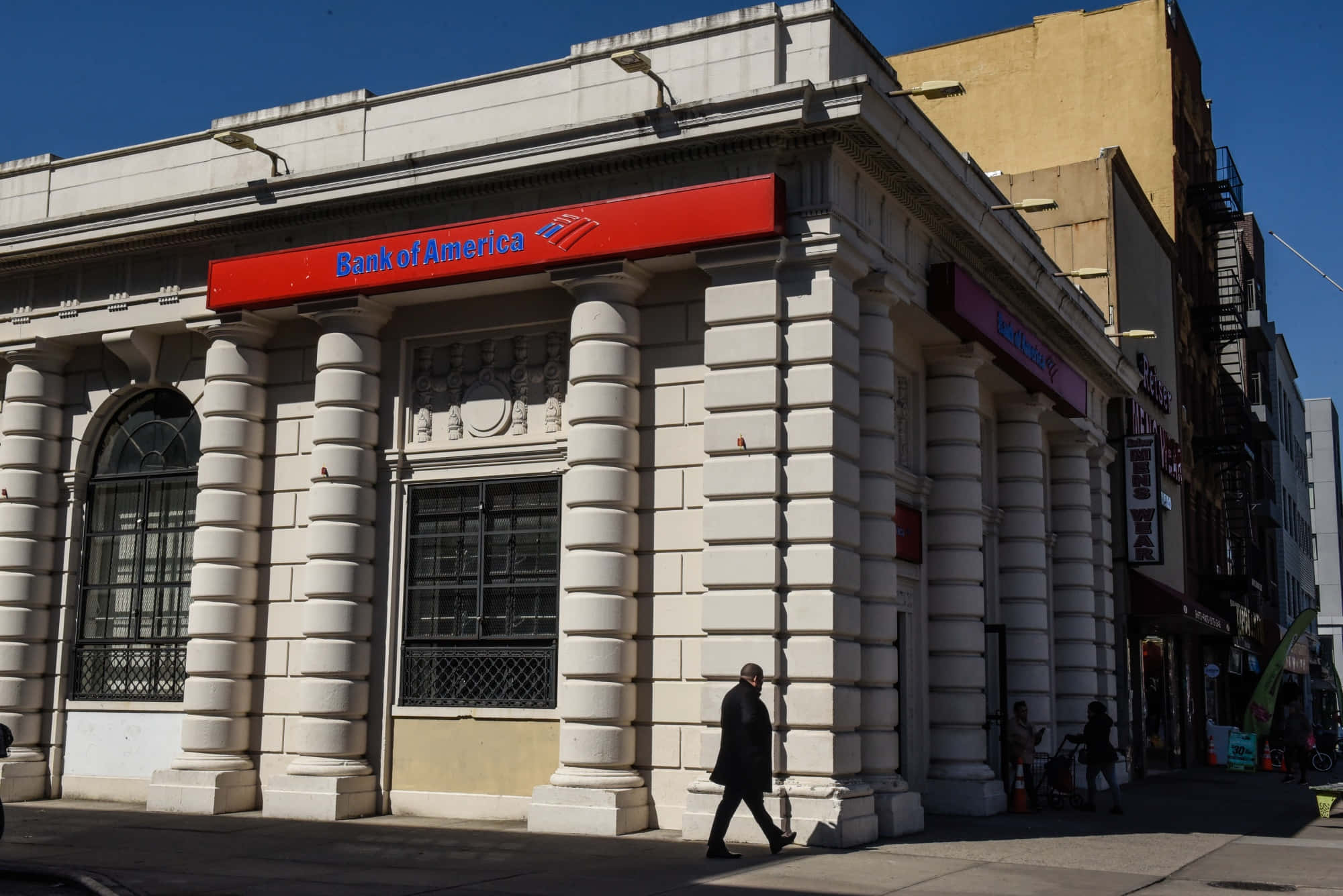 Bank 桌布 壁紙 — 一座白色石造的美國銀行大樓，擁有古典柱式，矗立在晴朗的藍天下，一名男子在陽光明媚的城市人行道上走過。