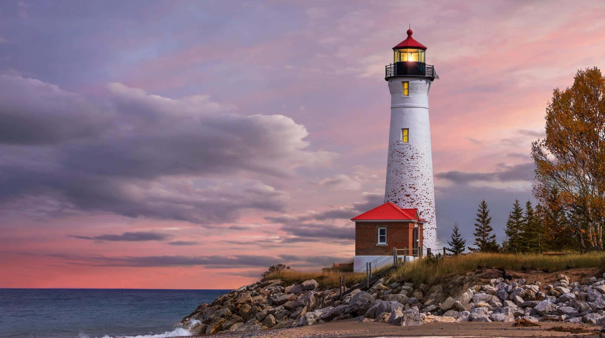 Lighthouse 桌布 壁紙 — 一座白色紅頂燈塔在岩石海岸線上，在充滿活力的粉紅色和紫色日落天空下溫暖地發光。