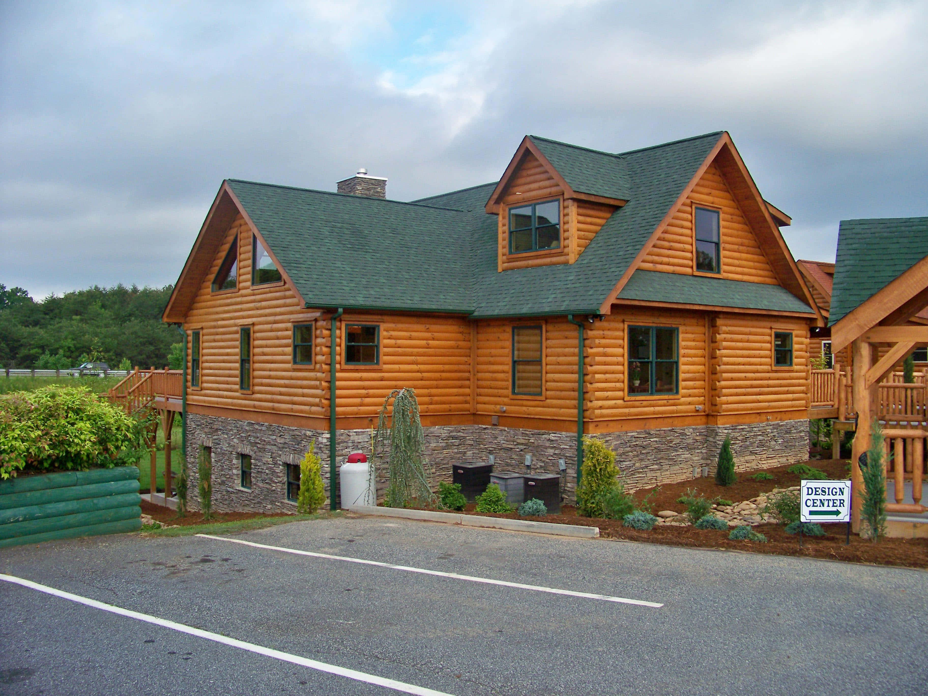 Log Cabin 桌布 壁紙 — 一座石地基、綠色屋頂的質樸木屋，矗立在多雲的天空下，旁邊是鋪砌的停車場。
