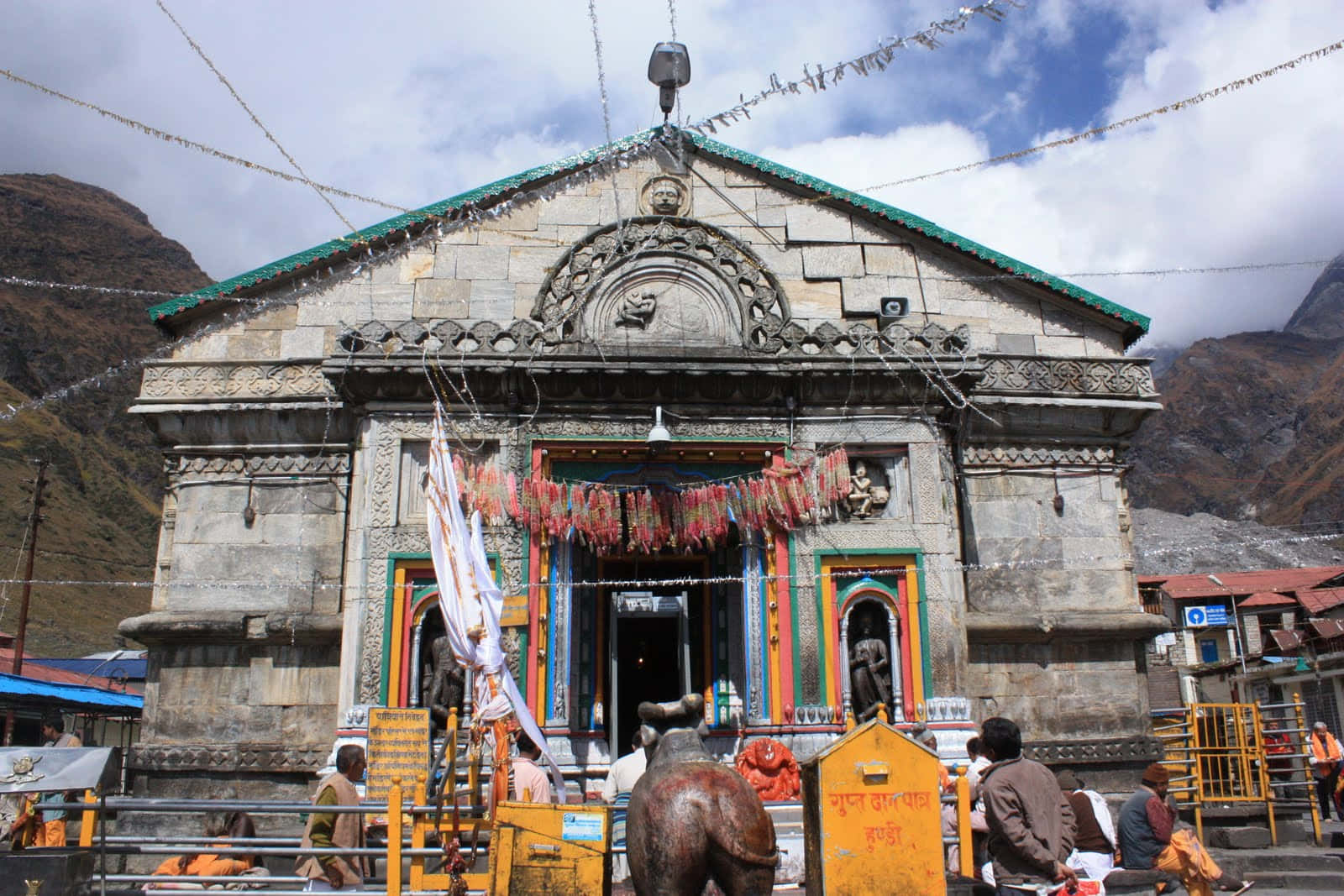 Kedarnath 桌布 壁紙 — 一座石廟，掛滿色彩繽紛的經幡，雕刻精緻，坐落在多雲的天空下的山谷中。