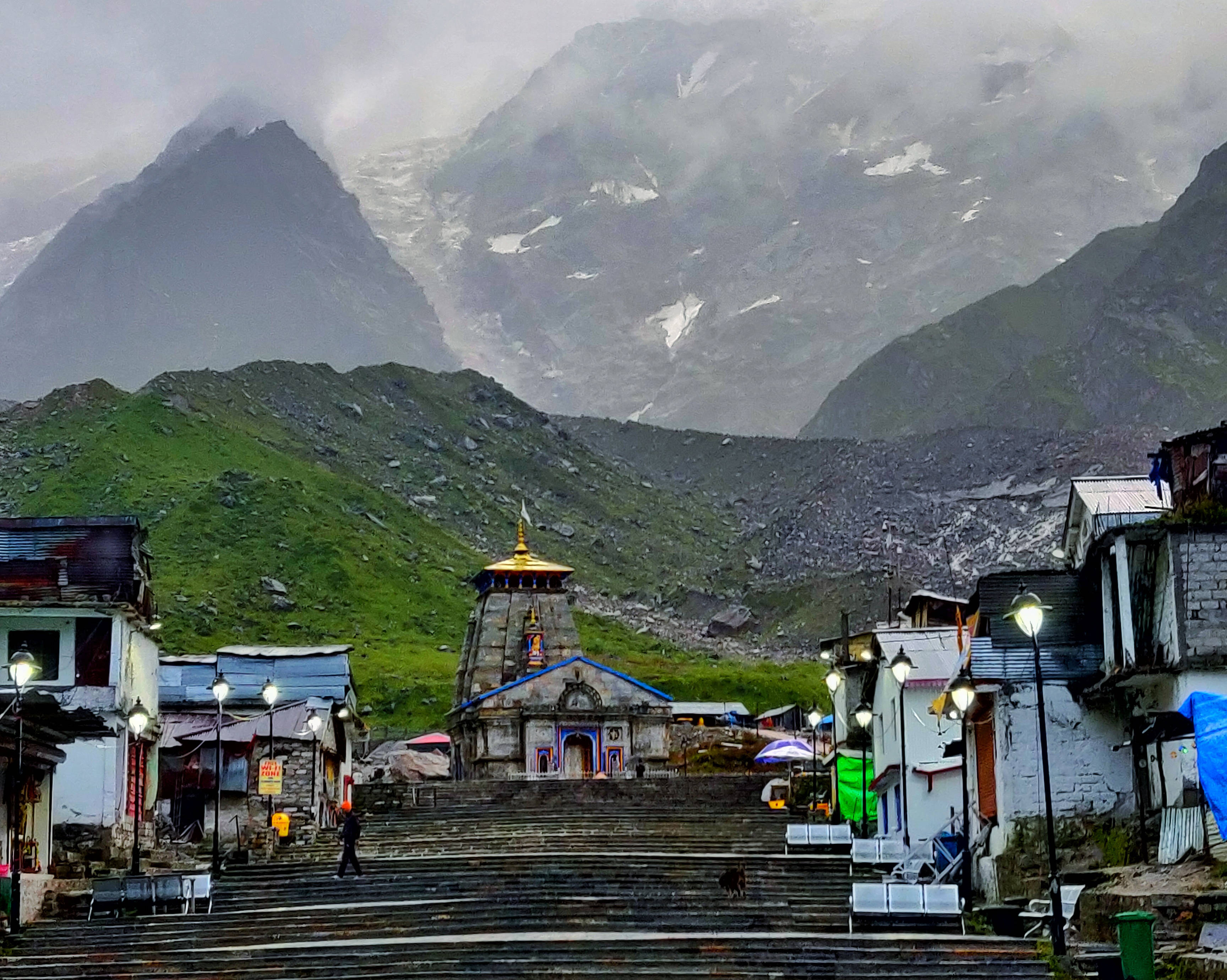 Kedarnath 桌布 壁紙 — 一座石廟帶有金色圓頂，坐落在山村的寬闊台階上，被霧氣繚繞的雪峰和綠色山坡環繞。