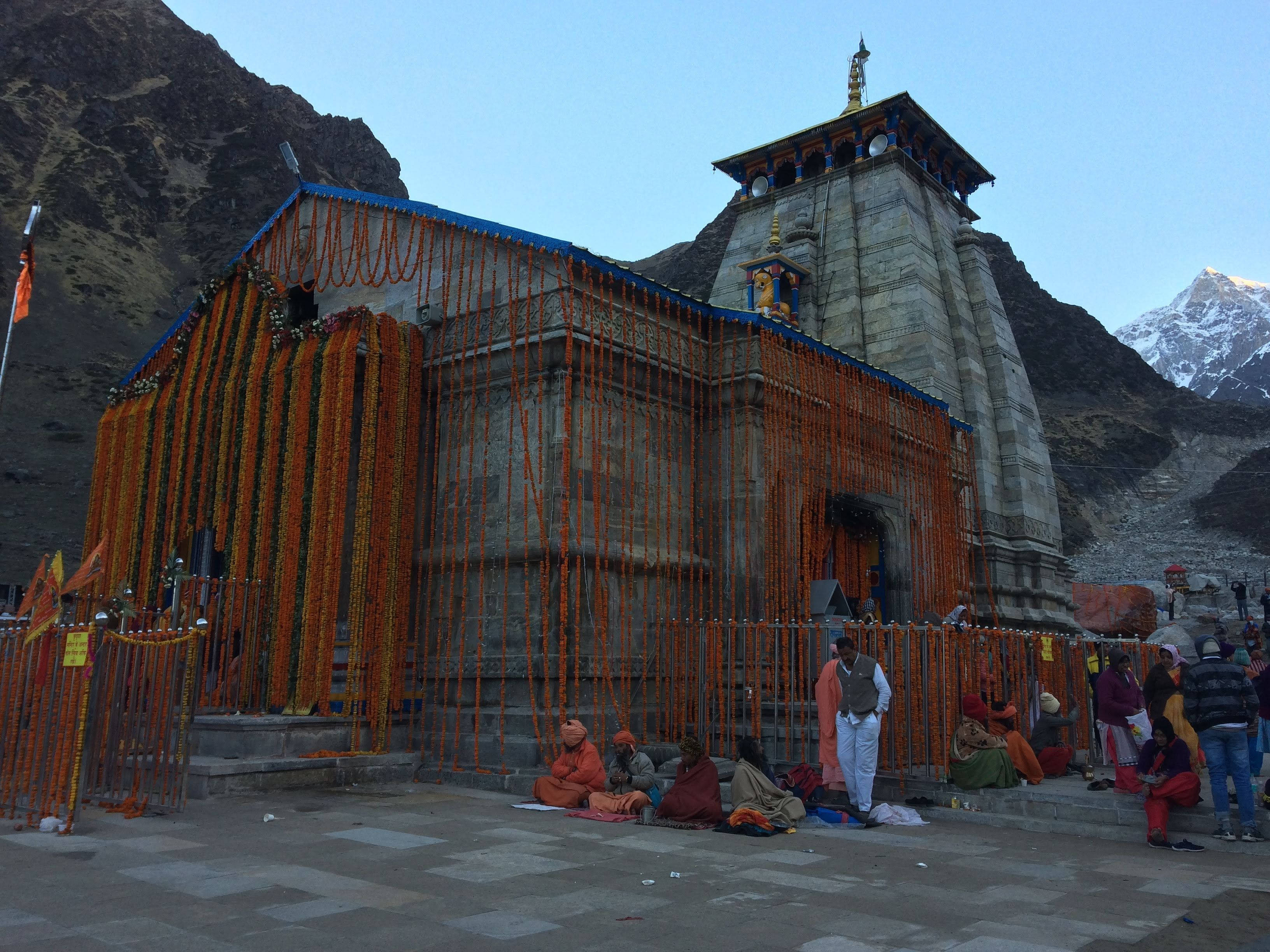 Kedarnath 桌布 壁紙 — 一座石廟掛滿鮮豔的橙色萬壽菊花環，依偎在崎嶇的山脈中，天空清澈湛藍。