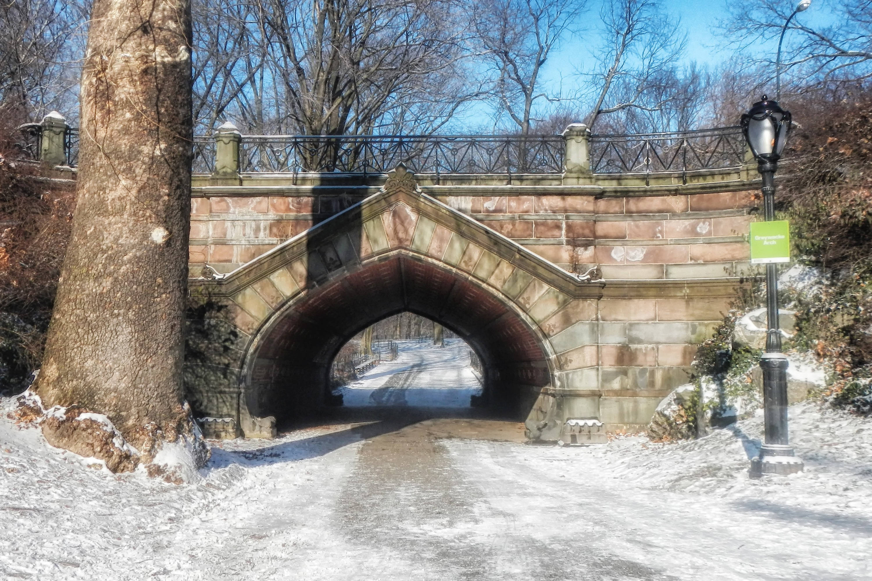 雪 桌布 壁紙 — 一座石拱橋，位於白雪覆蓋的公園中，有光禿禿的樹木和路燈，背景是清澈的藍天。