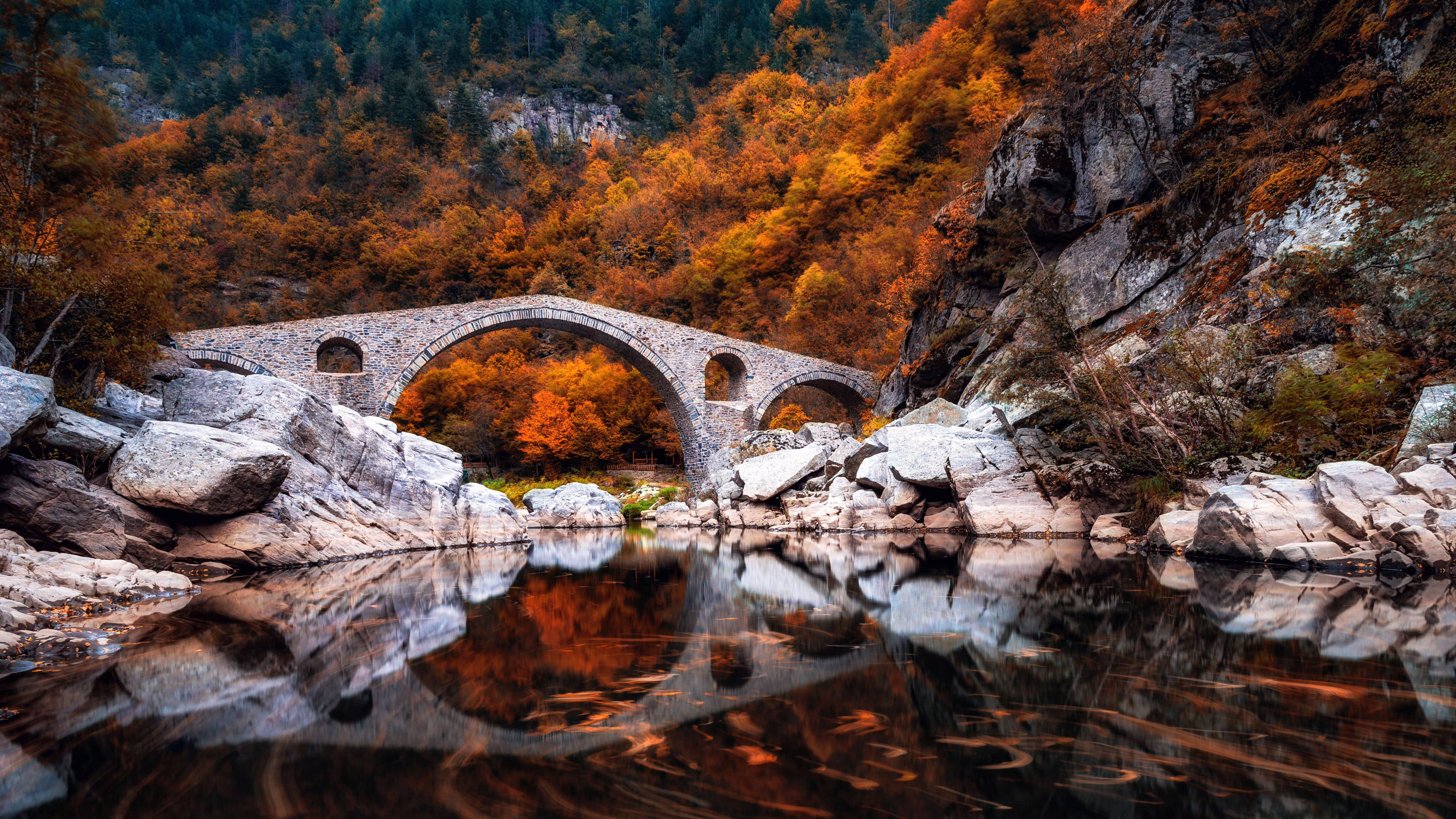 Bulgaria 桌布 壁紙 — 一座石拱橋橫跨平靜的河流，倒映著火紅的秋葉和崎嶇的懸崖，在柔和的光線下。
