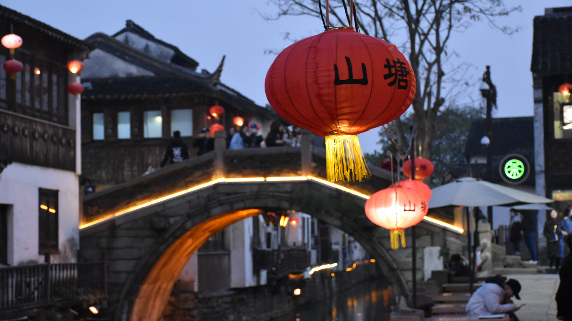 Suzhou 桌布 壁紙 — 一座石拱橋橫跨運河，在黃昏時分被掛在繁華傳統街道上方、帶有中文字樣的紅色發光燈籠照亮。