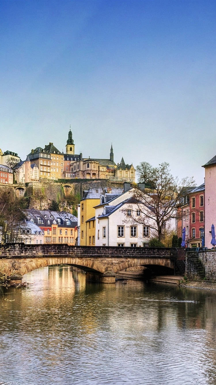 Luxembourg 桌布 壁紙 — 一座石橋在晴朗的藍天下拱跨平靜的河流，背景的山坡上點綴著色彩繽紛的歷史建築和一座城堡。