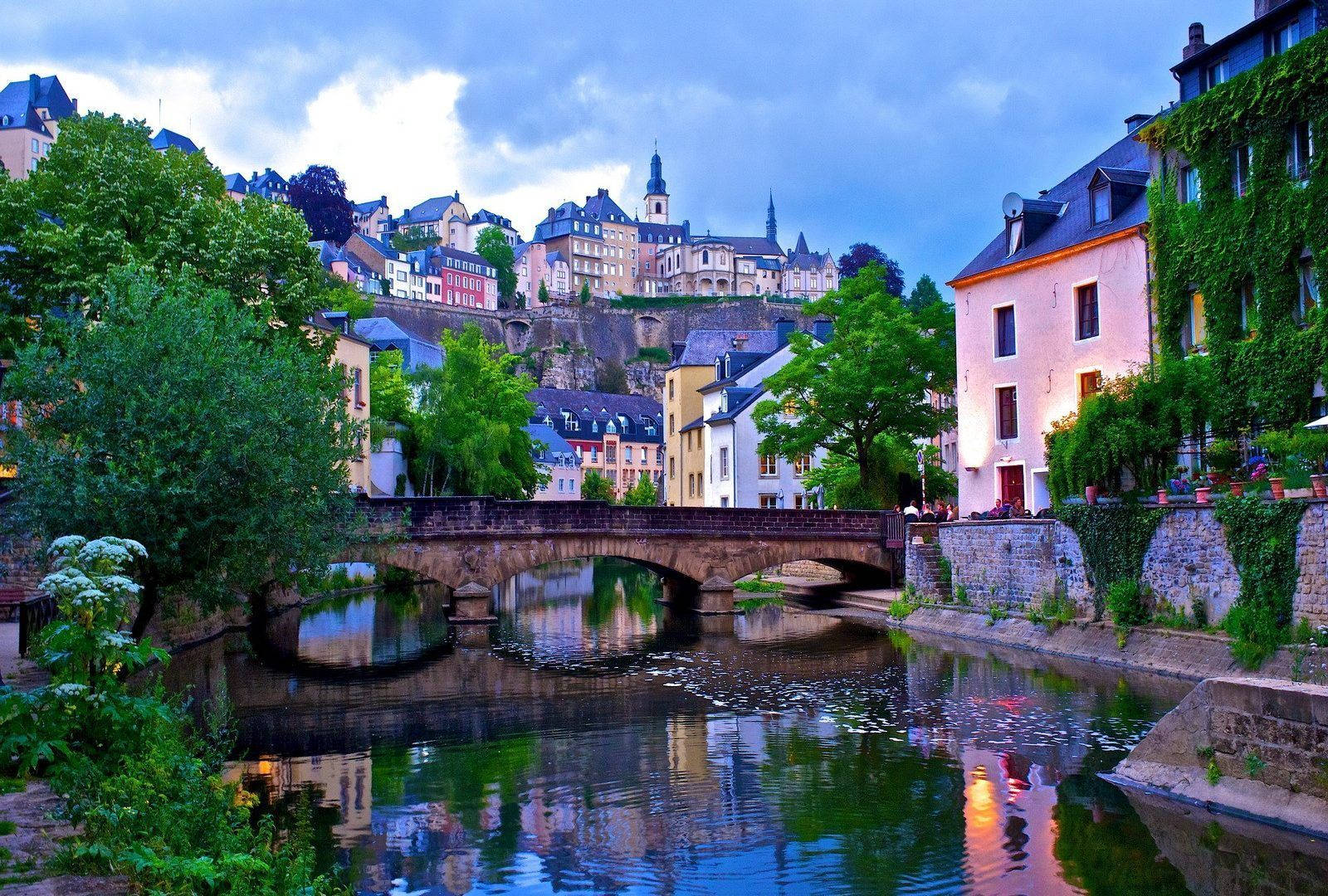 Luxembourg 桌布 壁紙 — 一座石橋拱跨平靜的河流，倒映著色彩繽紛的建築物和茂密的綠樹，在暮色天空下。