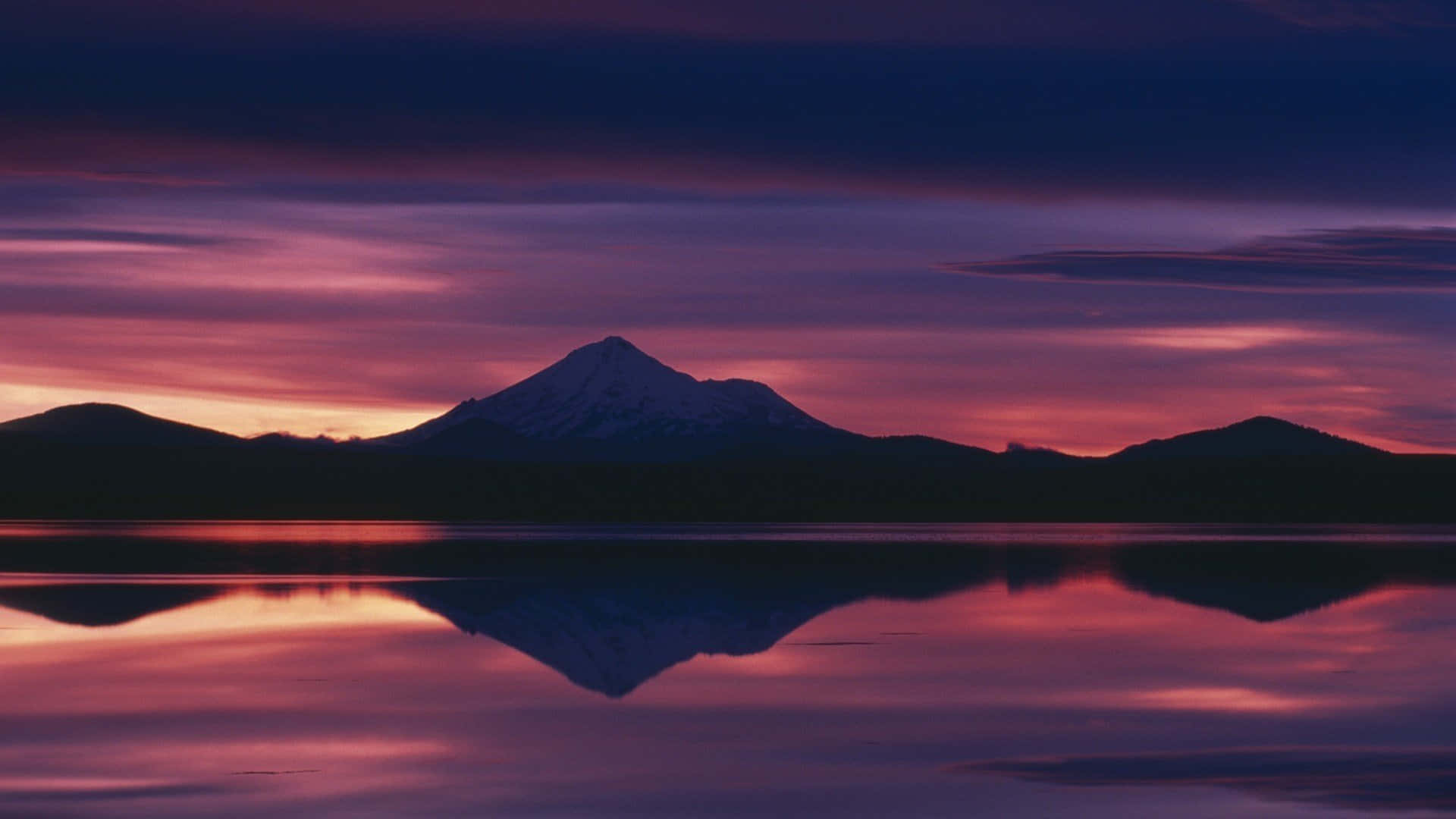 Sunset Aesthetic 桌布 壁紙 — 一座積雪的山峰在充滿活力的紫色和粉紅色日落映襯下形成剪影，完美倒映在下方平靜的湖面上。