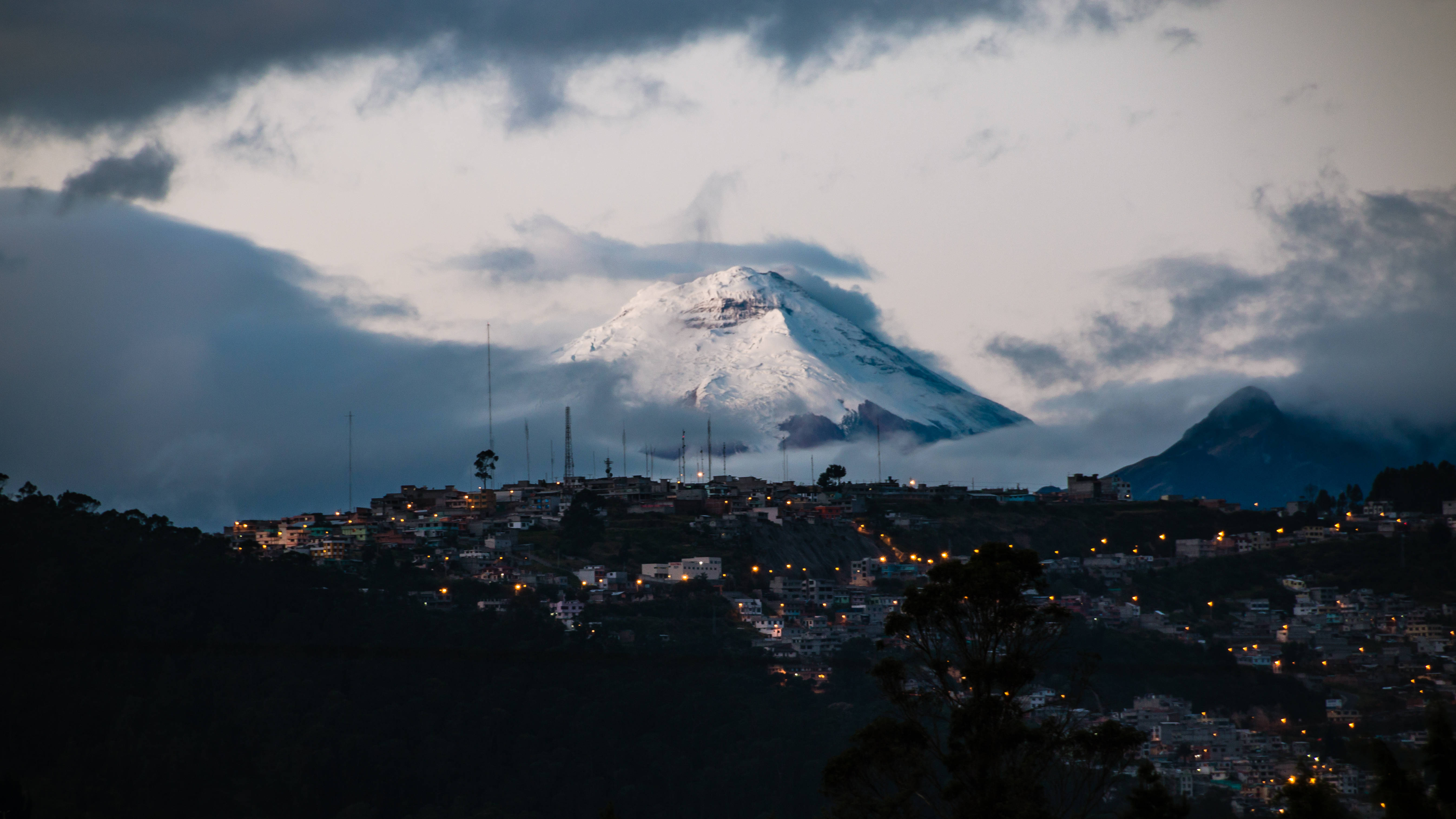 Ecuador 桌布 壁紙 — 一座積雪的山峰聳立於山丘小鎮之上，小鎮在多雲的暮色天空下閃爍著溫暖的燈光。