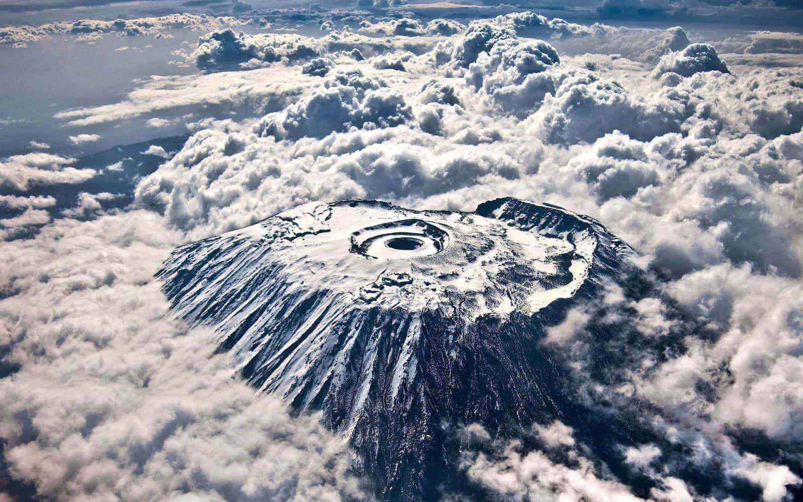 山脈 桌布 壁紙 — 一座積雪覆蓋的火山，帶有明顯的火山口，雄偉地聳立在蓬鬆的白雲海洋之上。