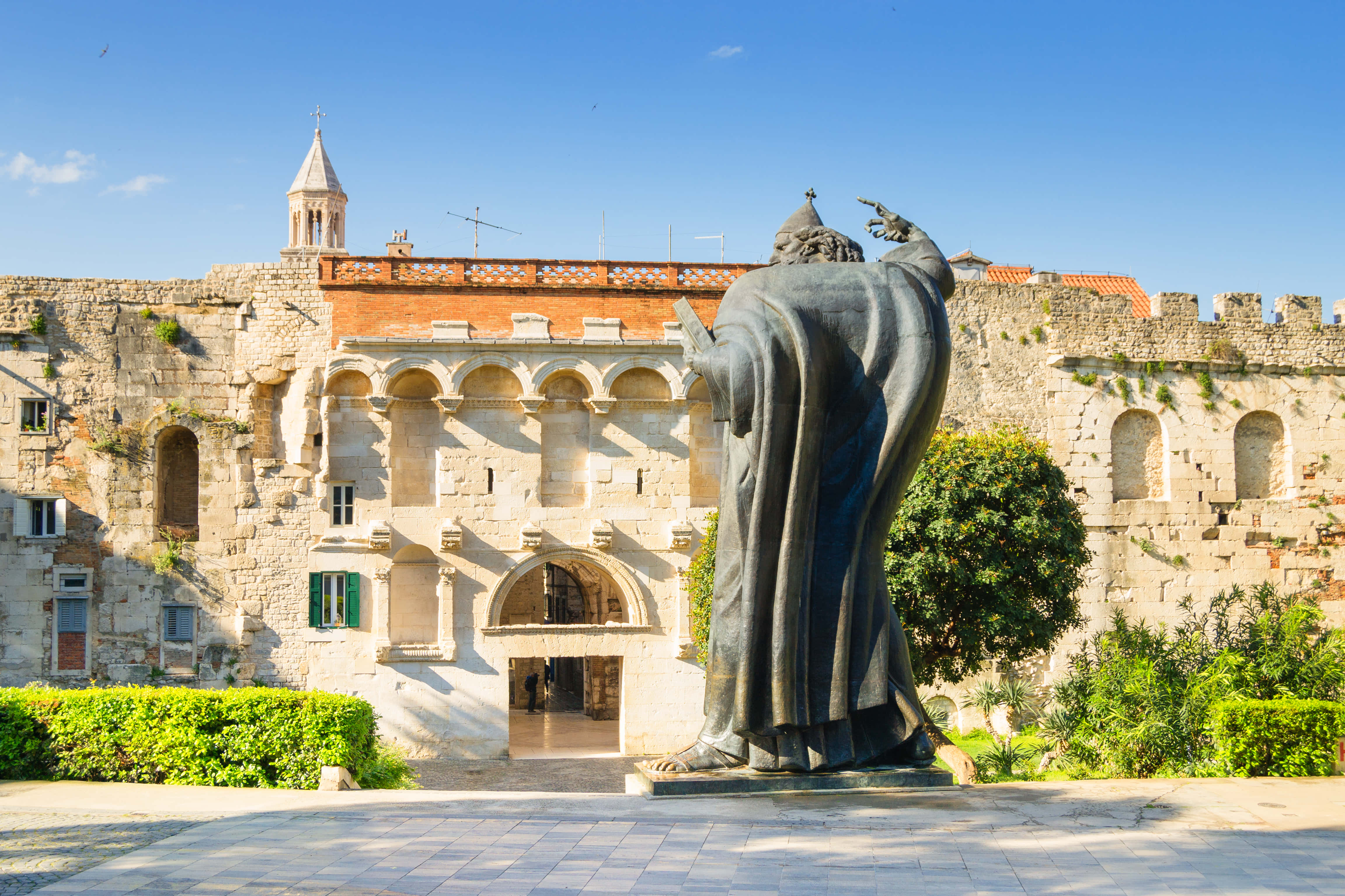 Croatia 桌布 壁紙 — 一座穿著長袍人物的銅像矗立在一座歷史悠久的石砌建築前，建築有拱形窗戶和鐘樓，背景是清澈的藍天。