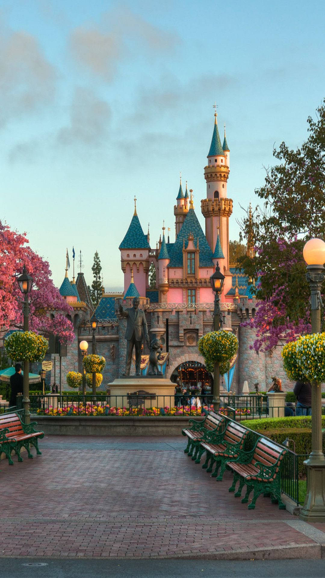 Disneyland Castle 桌布 壁紙 — 一座童話城堡，擁有藍色屋頂和粉紅色牆壁，在黃昏時分雄偉地矗立，周圍環繞著盛開的樹木和華麗的長椅。