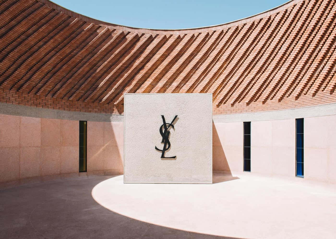 Ysl 桌布 壁紙 — 一座簡約現代建築，擁有弧形陶土屋頂，在陽光普照的庭院中投射出幾何陰影，庭院中有一個突出的YSL標誌。
