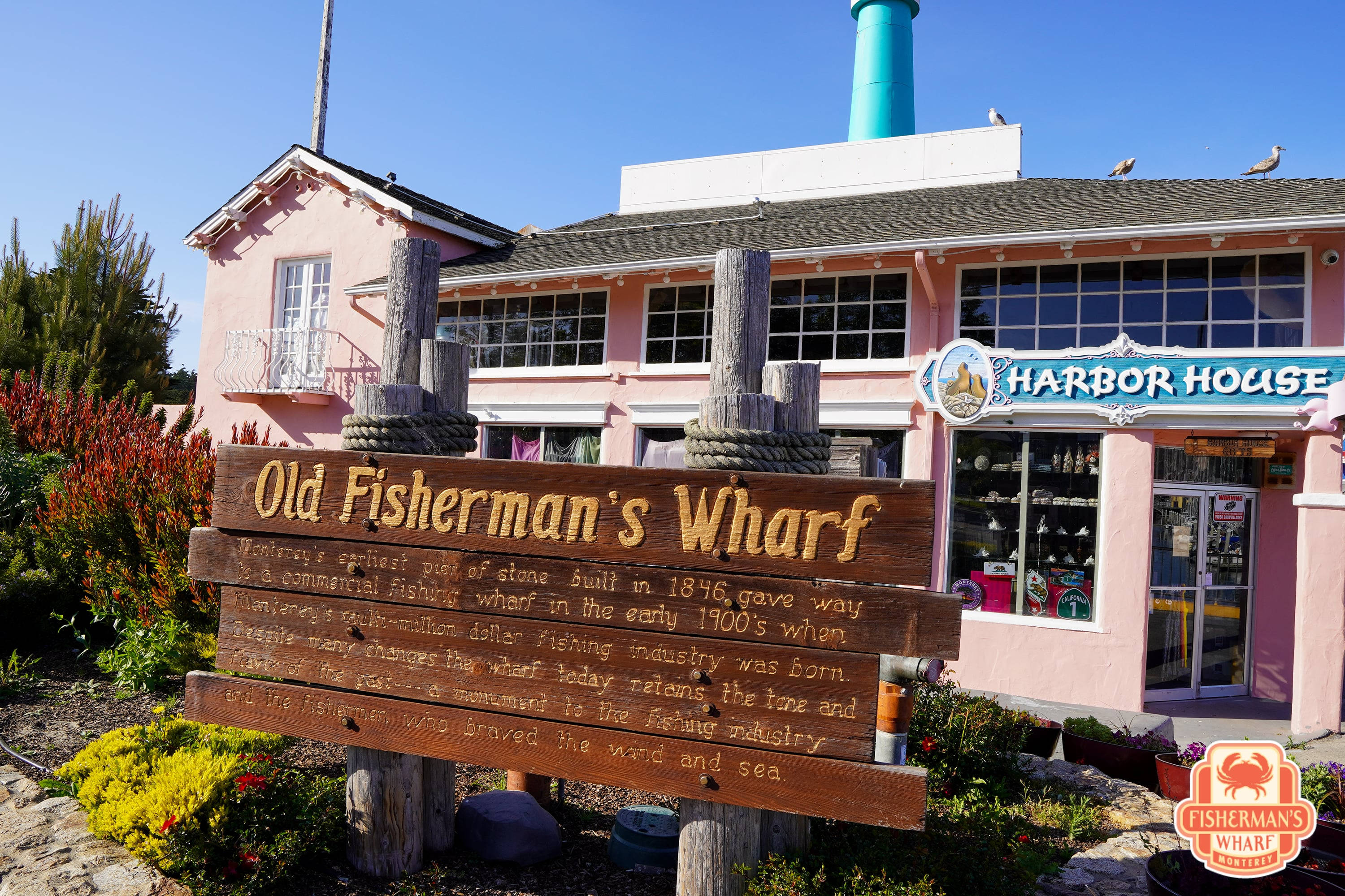 加州 桌布 壁紙 — 一座粉紅色的Harbor House建築，帶有Old Fisherman's Wharf的木製招牌，矗立在晴朗的藍天下，周圍有茂密的綠植和一個青綠色的煙囪。