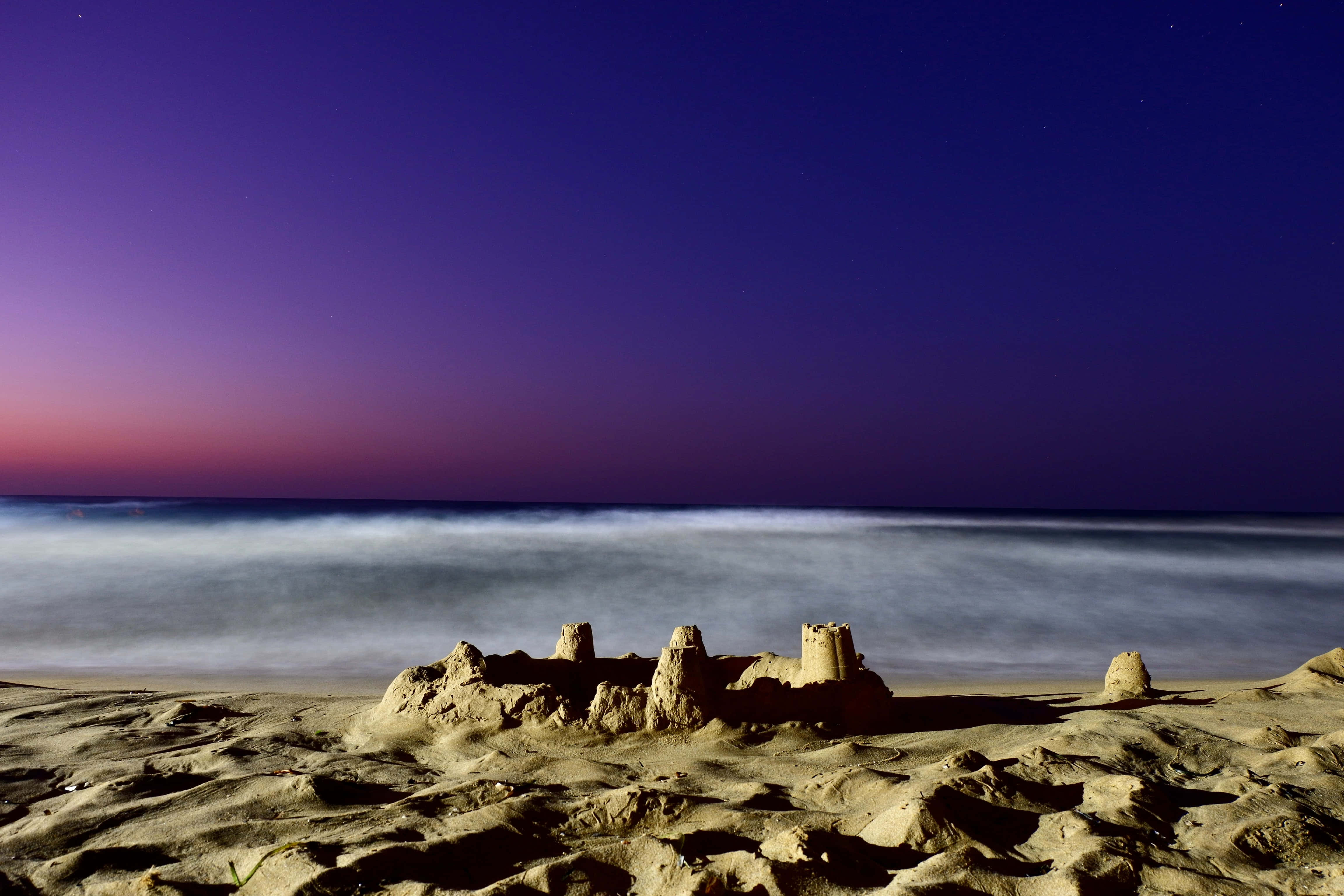 Sandcastles 桌布 壁紙 — 一座精緻的沙堡矗立在沙灘上，背景是染上紫色和粉紅色調的暮色天空，海洋在背景中輕輕流動。
