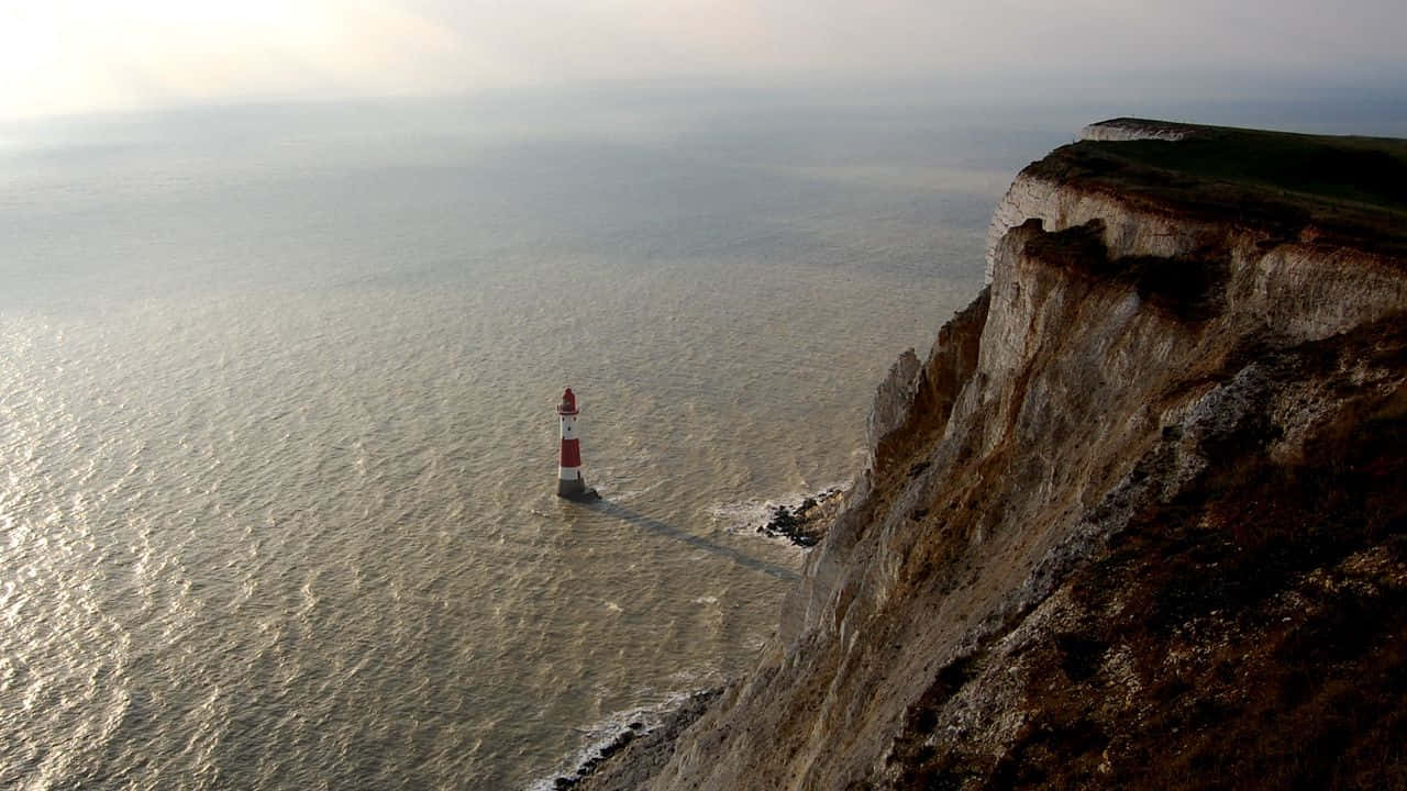 Cliff 桌布 壁紙 — 一座紅白條紋燈塔孤獨地矗立在海上岩石露頭旁，旁邊是霧氣朦朧的天空下高聳的白色懸崖。