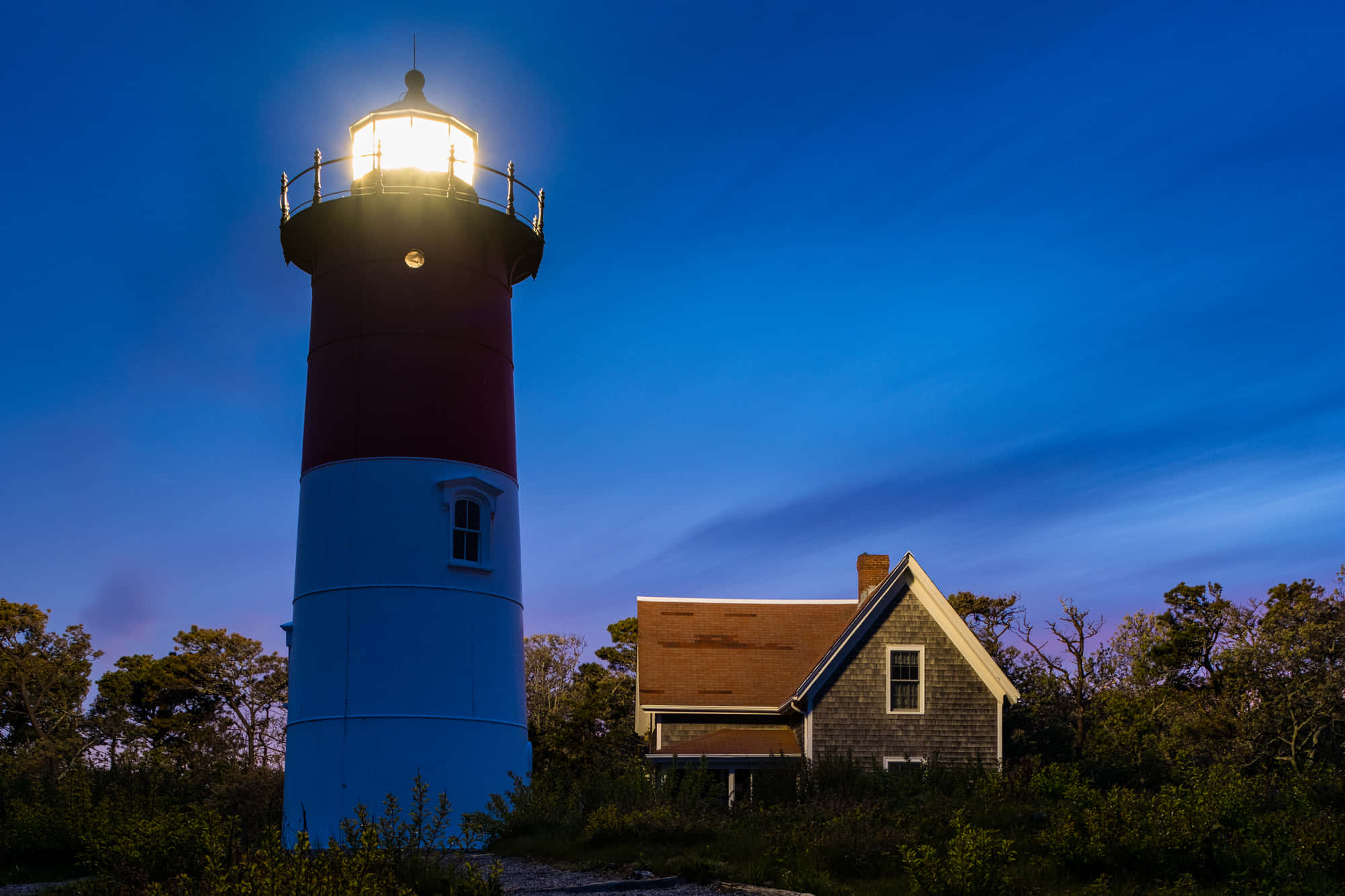 Lighthouse 桌布 壁紙 — 一座紅白燈塔在深藍色的暮色天空中發出光芒，旁邊有一座小小的看守人小屋。