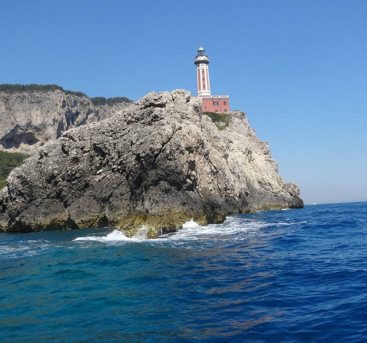 Capri 桌布 壁紙 — 一座紅白燈塔棲息在崎嶇的懸崖上，俯瞰著清澈天空下深藍色的海浪。