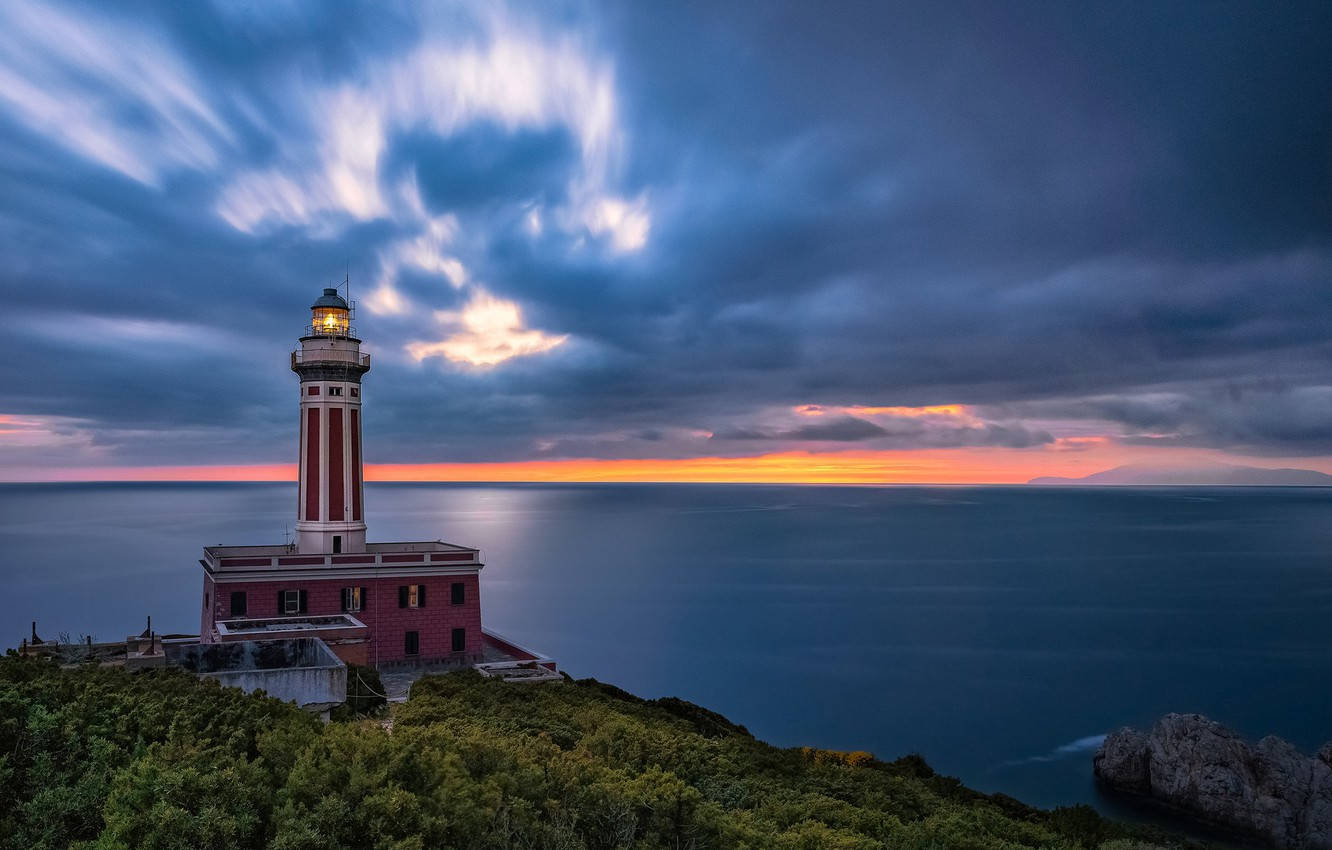 Capri 桌布 壁紙 — 一座紅白燈塔矗立在懸崖上，夕陽西下，天空有條紋雲彩，下方是平靜的海面。