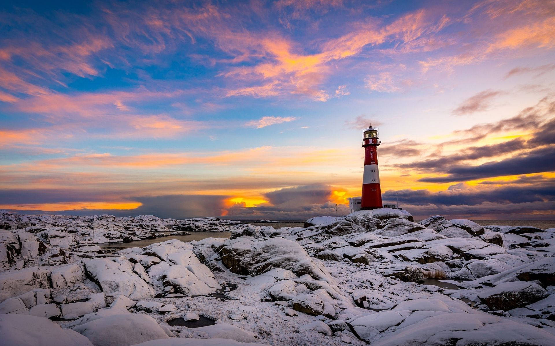 Lighthouse 桌布 壁紙 — 一座紅白燈塔矗立在積雪覆蓋的岩石上，天空呈現出橙色、粉紅色和藍色的絢麗日落色彩。