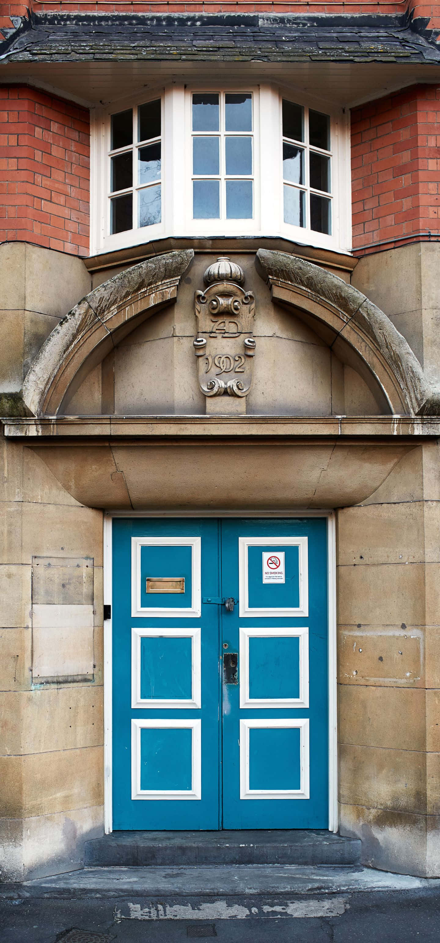 1440x3088 桌布 壁紙 — 一座紅磚建築的凸窗下方，風化石拱門內有一扇帶白色飾邊的鮮豔藍綠色雙門。