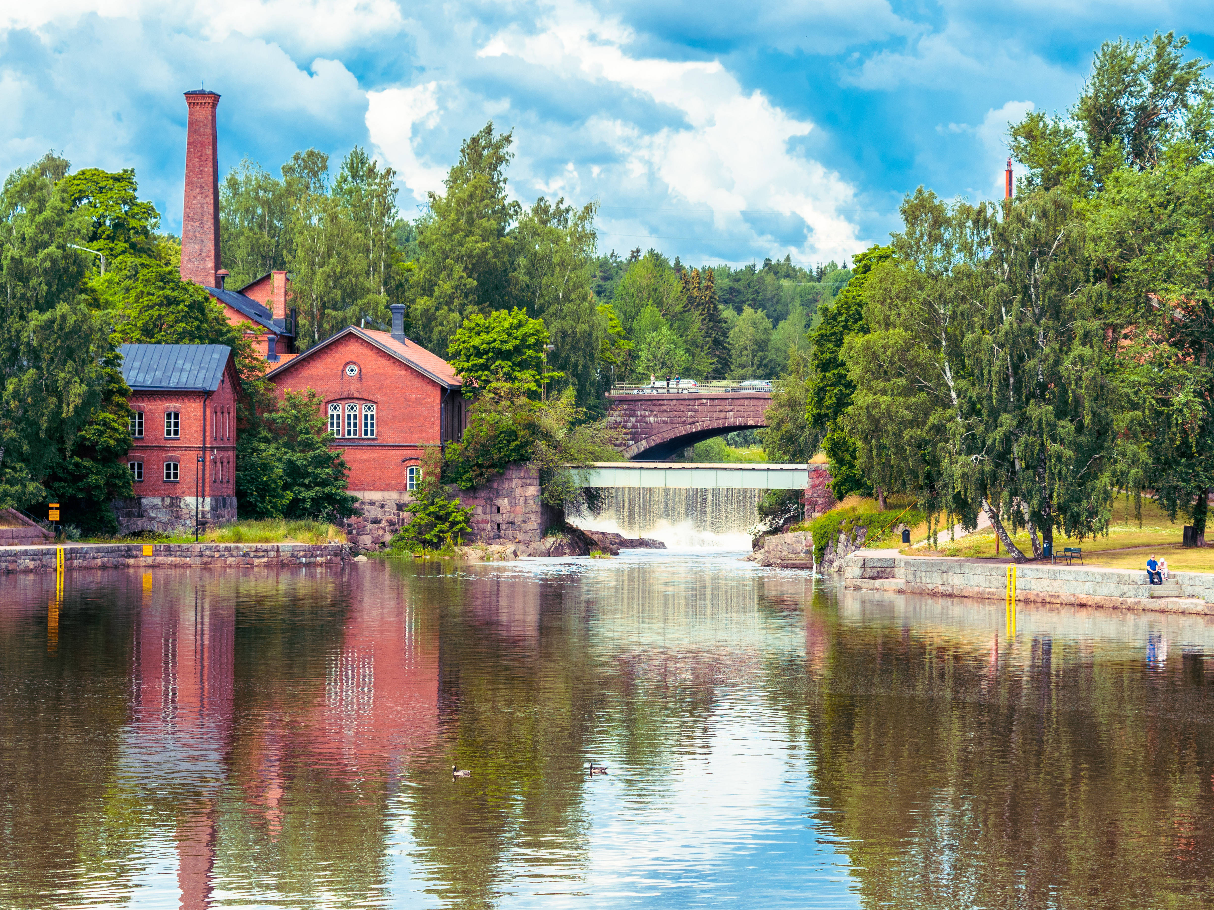 Finland 桌布 壁紙 — 一座紅磚磨坊，帶有高煙囪，矗立在河邊，河上有石橋和瀑布，天空湛藍，白雲朵朵。