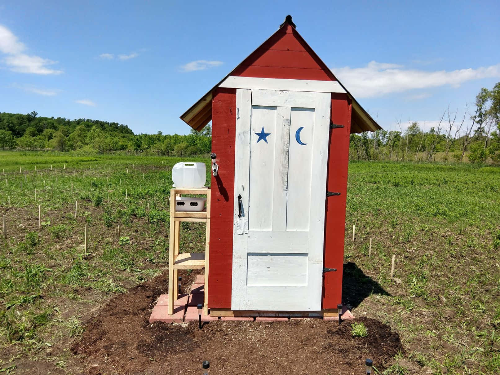 Outhouse 桌布 壁紙 — 一座紅色木製戶外廁所，帶有白色門，門上有藍色星星和新月圖案，矗立在晴朗藍天下的綠色田野中。