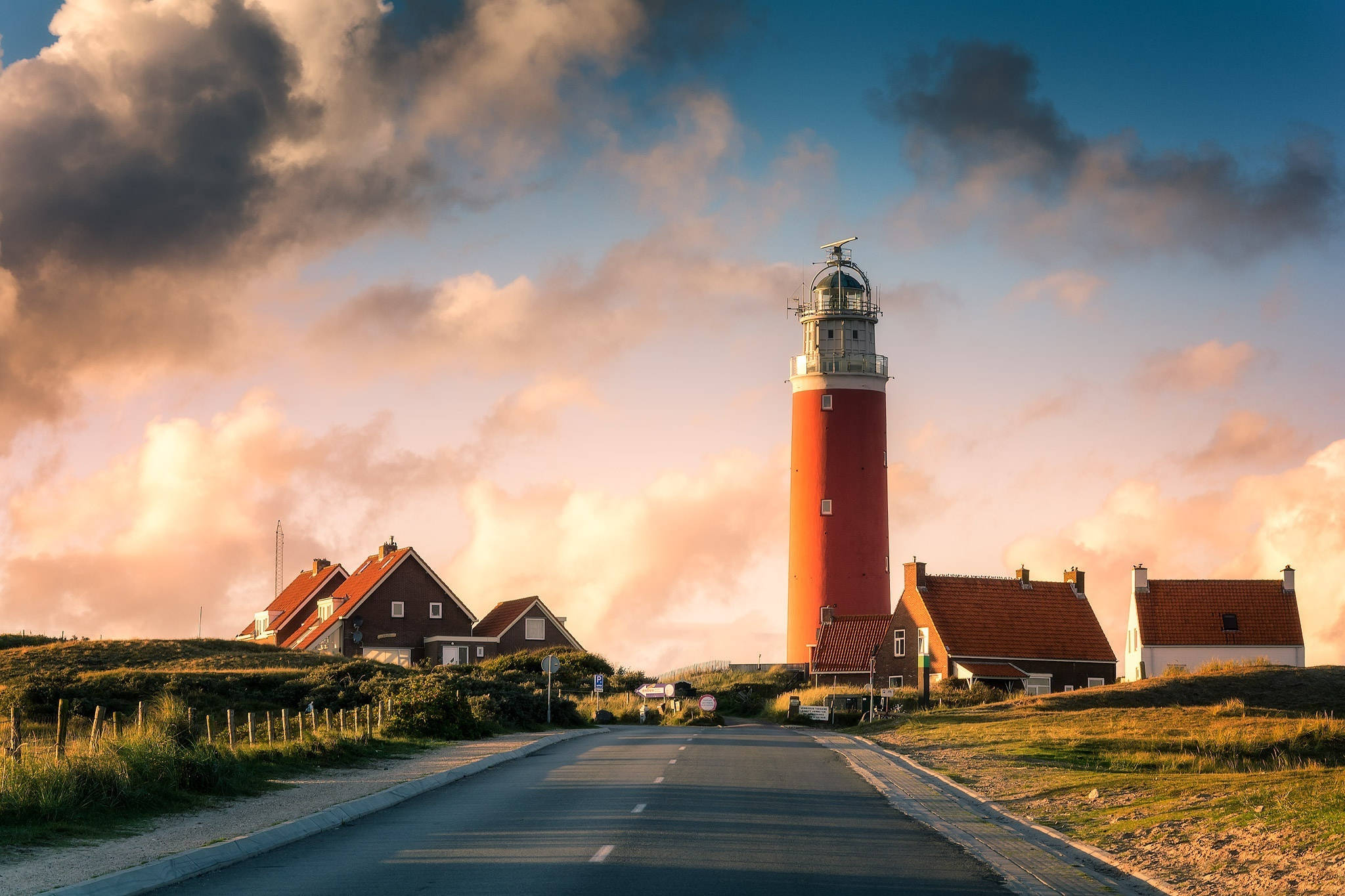 Netherlands 桌布 壁紙 — 一座紅色燈塔高聳在古雅房屋旁，一條道路在戲劇性的日落天空下通向它。