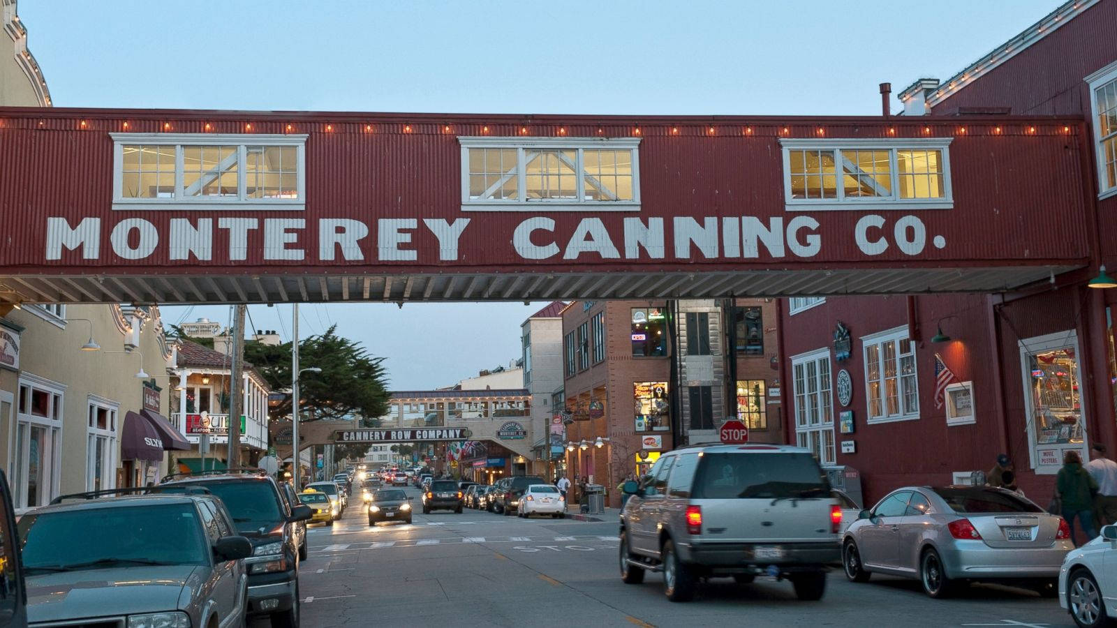 Building 桌布 壁紙 — 一座紅色的 Monterey Canning Co. 行人天橋橫跨一條繁忙的街道，街道上有汽車和商店門面，正值黃昏時分。