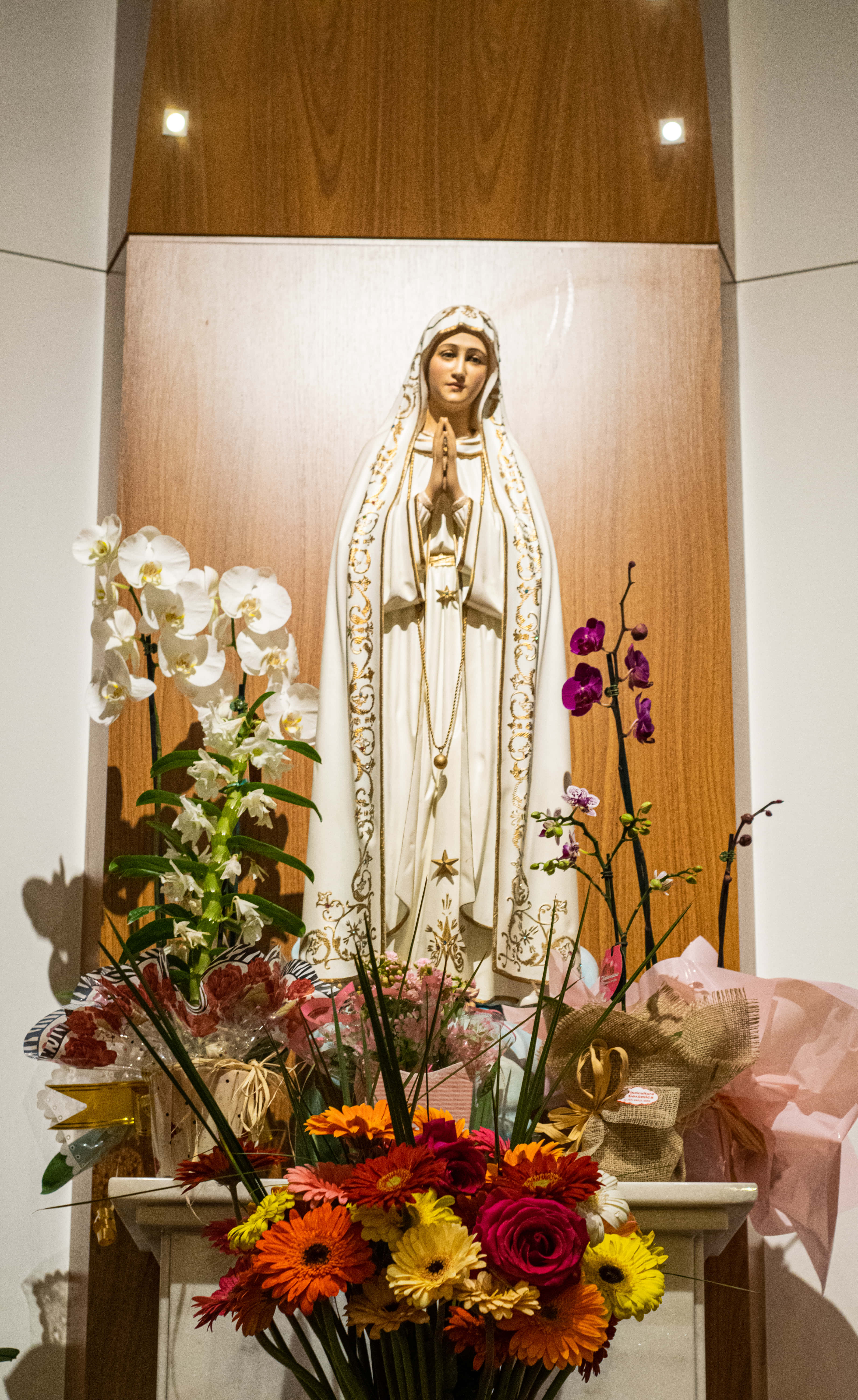 Mother Mary 桌布 壁紙 — 一座純白寧靜的聖母瑪利亞雕像，矗立在木製壁龕中，周圍環繞著鮮豔的花卉佈置。