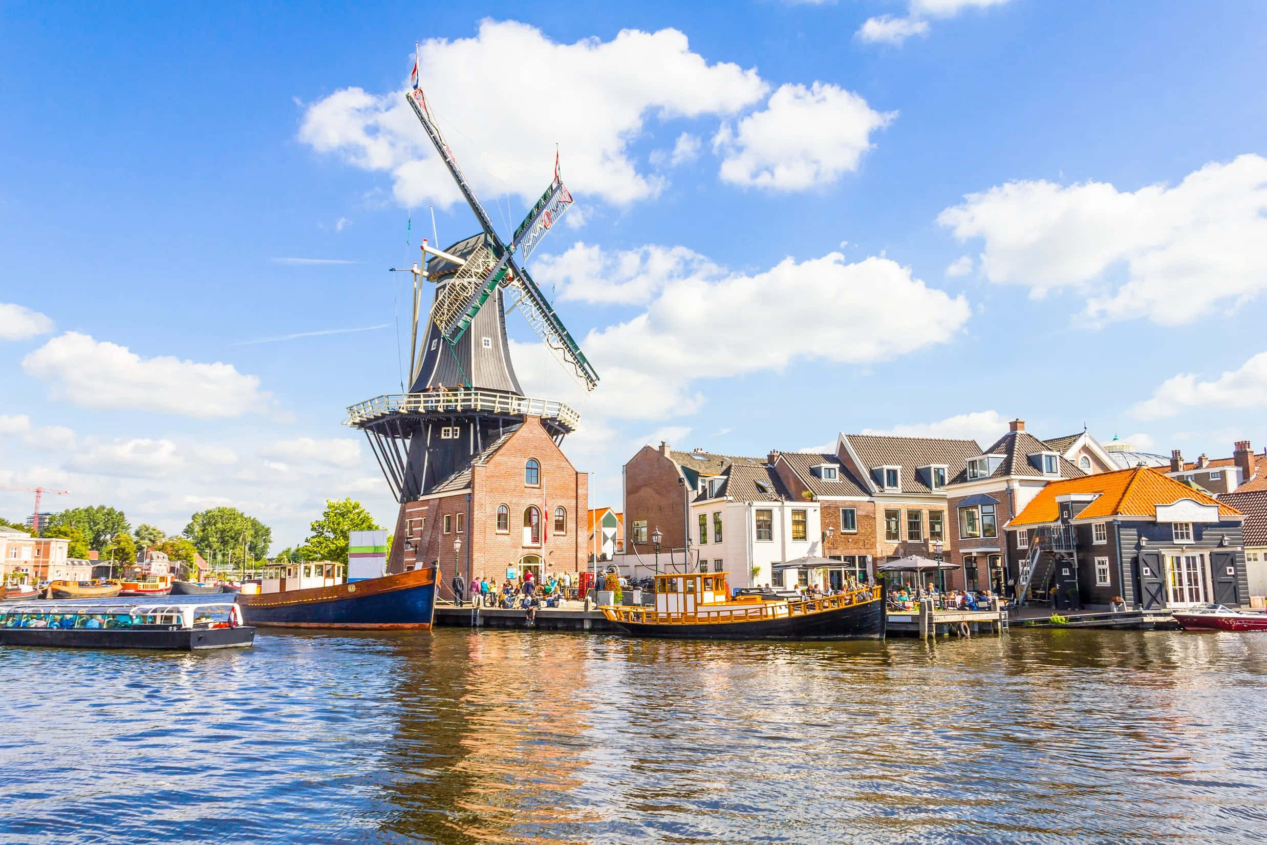 Netherlands 桌布 壁紙 — 一座經典的荷蘭風車聳立在運河旁，運河上有船隻和傳統房屋，上方是晴朗的藍天和白雲。