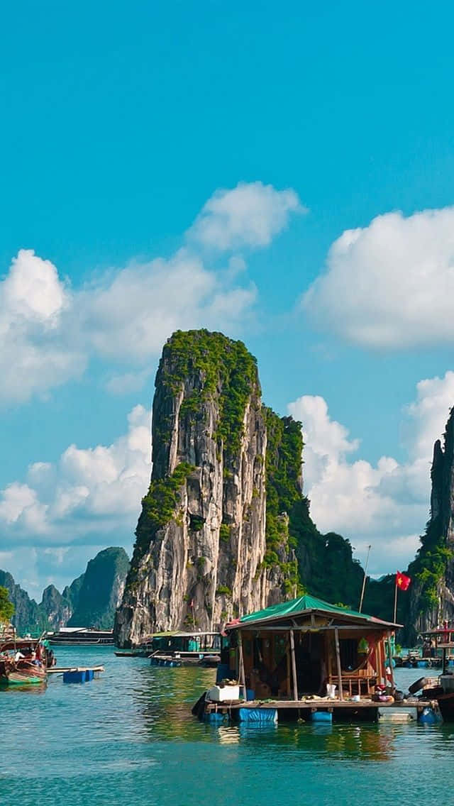 越南 桌布 壁紙 — 一座綠色屋頂的漂浮木屋坐落在平靜的碧綠水面上，下方是雄偉的石灰岩懸崖，覆蓋著茂密的綠色植物，上方是明亮的藍天和蓬鬆的白雲。